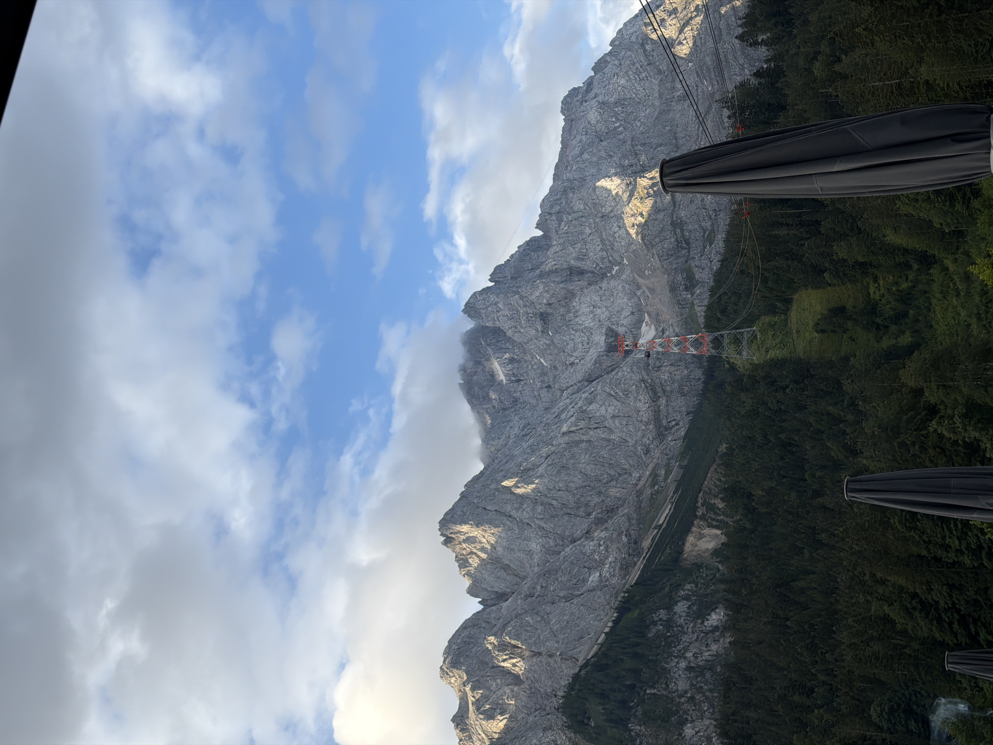 The image depicts a view of the Zugspitze, Germany's highest peak, from the vicinity of Grainau. In the foreground, several closed dark grey umbrellas are visible, suggesting an outdoor seating area. Behind them, a dense, dark green forest covers the lower slopes of the mountains. A ski lift pylon, red and white, stands prominently on the mountainside, with cables extending upwards. The rugged, grey rocky peaks of the Zugspitze dominate the background, partially obscured by clouds and illuminated by sunlight, casting shadows across their craggy surfaces. The sky is a mix of blue and white clouds, with patches of sunlight breaking through. The scene conveys a sense of majestic natural beauty combined with man-made infrastructure for tourism.