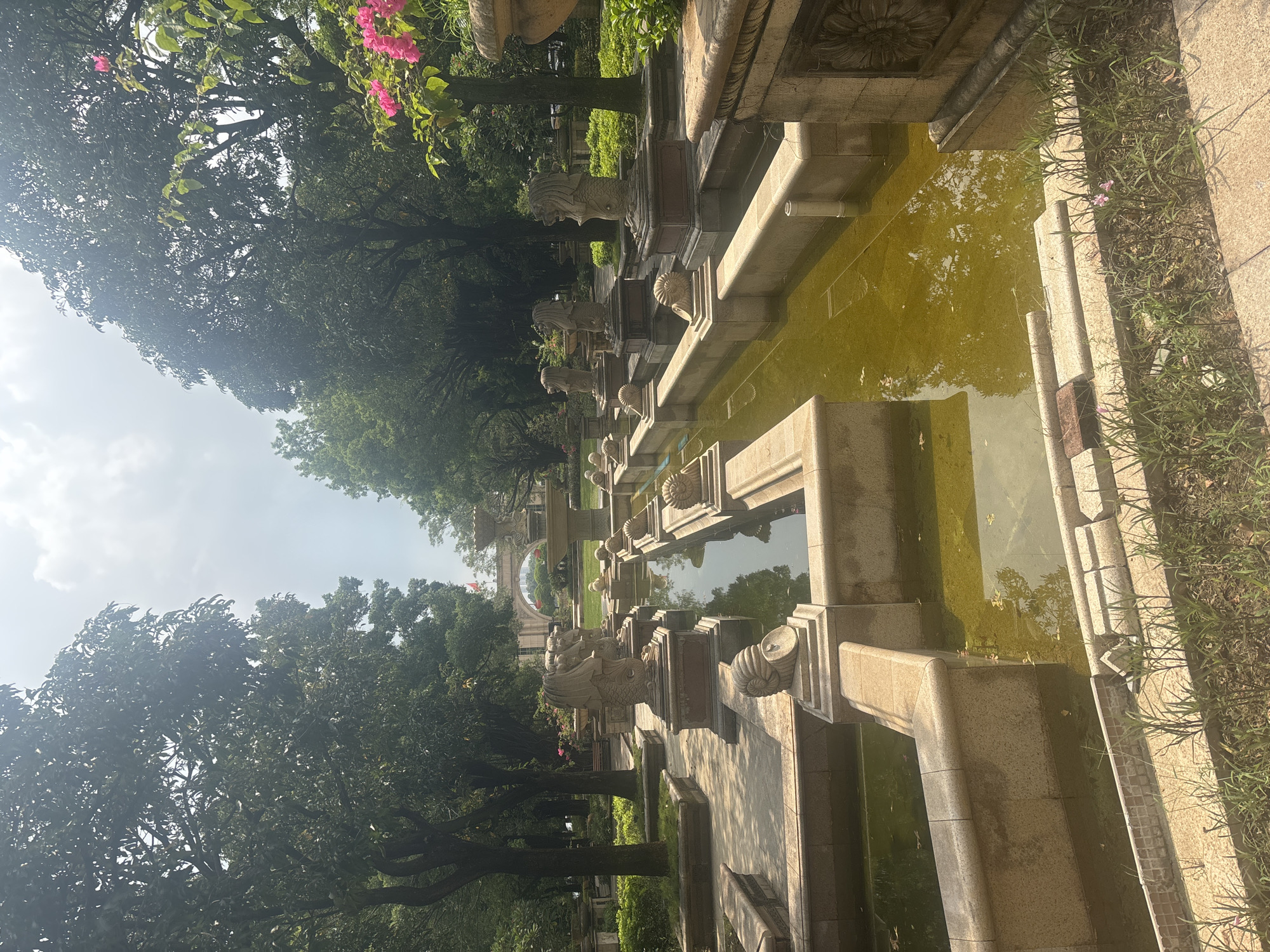 The image depicts a public park in Nanhai District, China, featuring a large, multi-level water fountain. The fountain is constructed from stone and appears to be aged, with some water algae visible in the pools. Ornate sculptures of mythical creatures, resembling mermaids or sphinxes with lion heads and fish tails, are strategically placed along the fountain's edges. These statues stand on stone pedestals. In the background, an archway with a structure resembling a building or monument is visible, with a flag atop it. Mature trees with dense foliage surround the fountain area, and the sky above is partly cloudy. The lighting suggests it is daytime, with the sun casting shadows on the stone pathways and structures. The overall impression is of a historical or ornamental garden design.