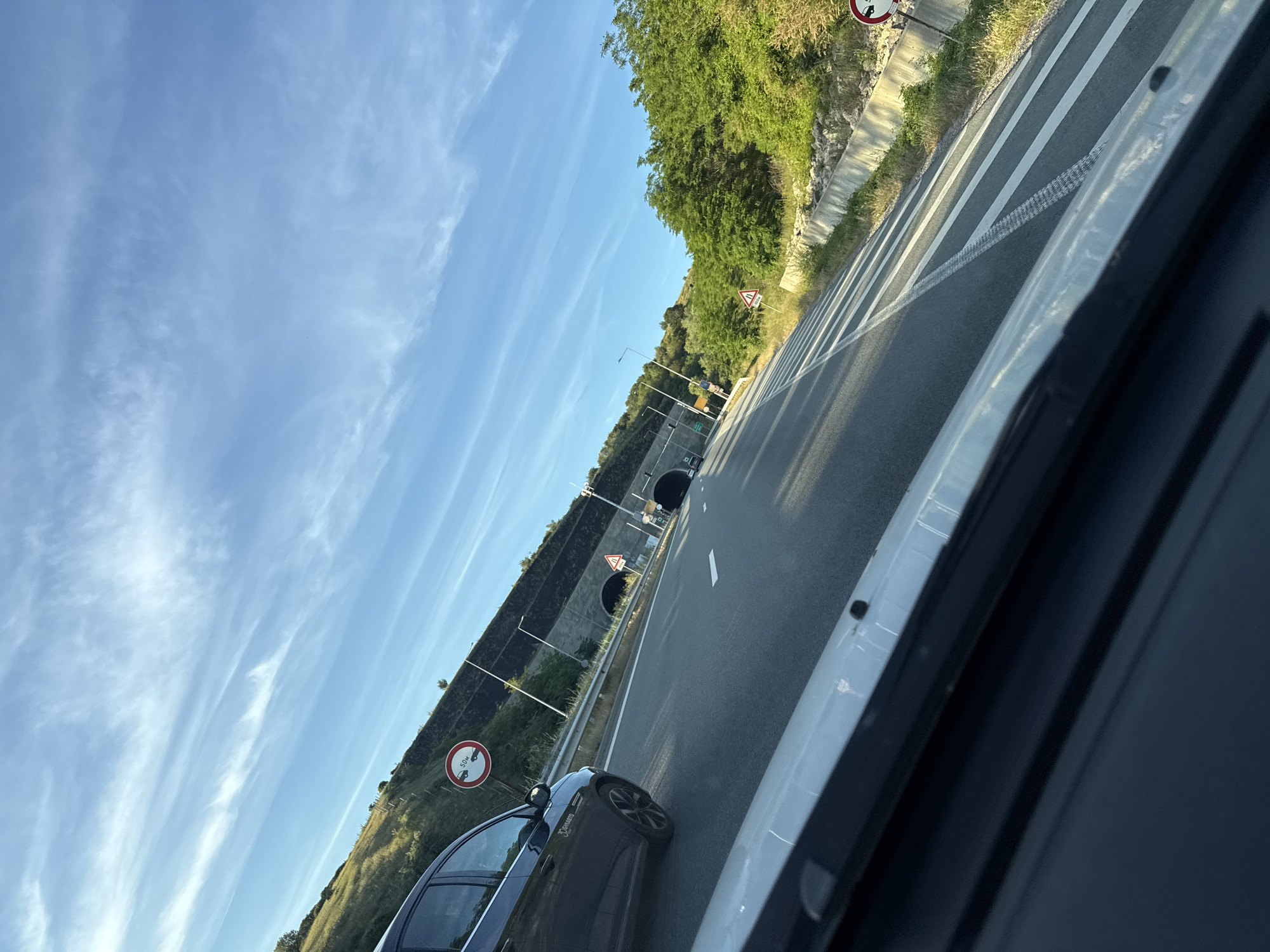 The image captures a perspective from within a vehicle traveling on a highway. The road ahead curves to the right, with multiple lanes marked by white lines. On the left side of the highway, a dark-colored car is visible in the adjacent lane. Beyond the car, a steep, grassy embankment rises, topped by a rock-faced wall and a tunnel entrance. A traffic sign with red and white circular markings and the text "50M" is positioned on the embankment. On the right side of the highway, a line of trees and vegetation borders the road, with a concrete barrier and more traffic signs visible. The sky is clear blue with scattered wispy clouds, illuminated by sunlight. The overall scene suggests a journey through a mountainous or hilly region, possibly a tunnel approach. Without specific landmarks or street signs, it is not possible to definitively confirm the location as Blatino, Bulgaria, though the scenery is consistent with such an area.