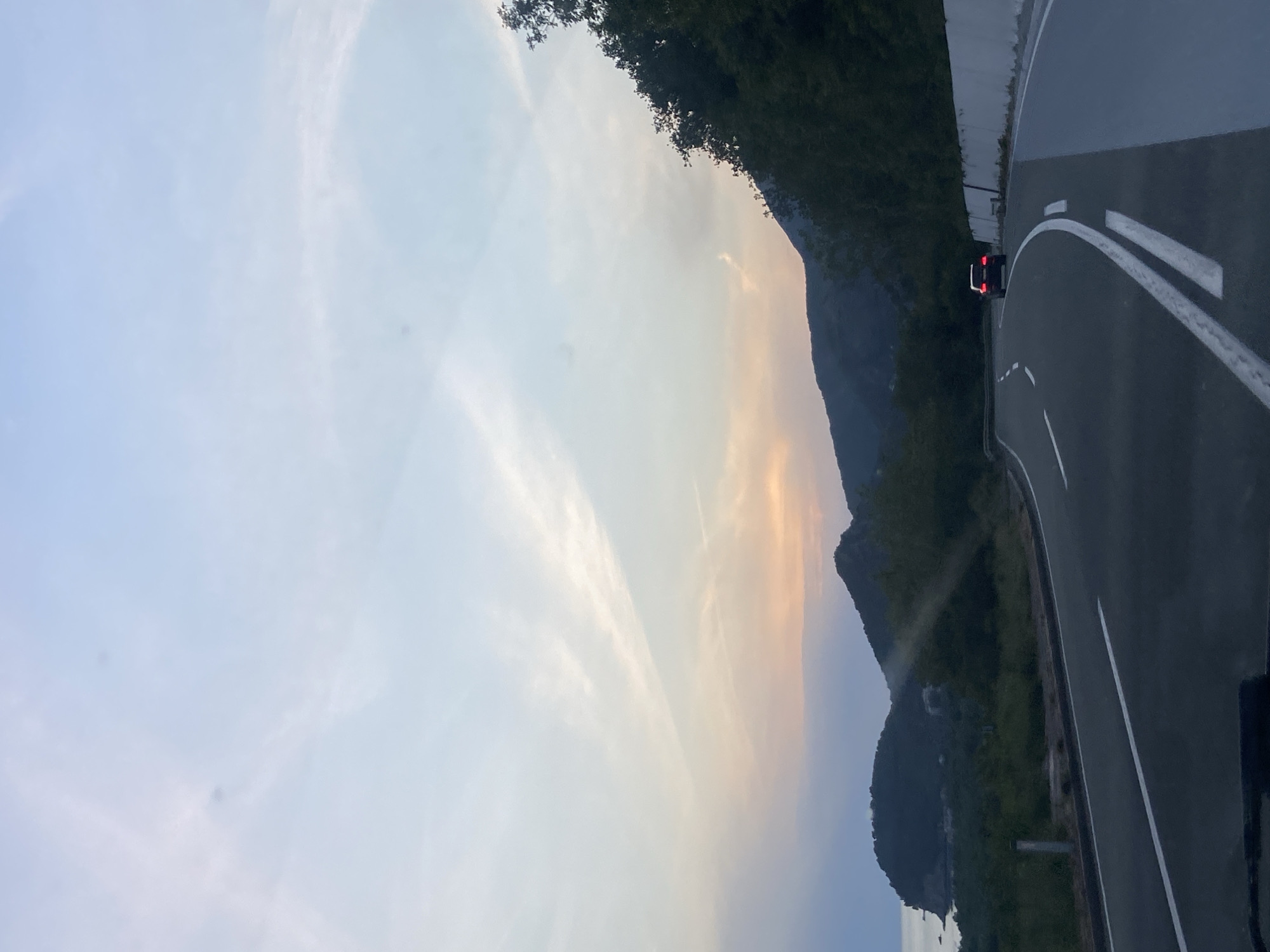 The image shows a view from a road in Golubovići, Montenegro. The road has white lane markings and a shoulder. A vehicle with illuminated tail lights is visible on the road. Lush green vegetation covers the mountainous terrain on the right side of the road. The sky is light blue with some wispy clouds, with a tinge of orange near the horizon. The Adriatic Sea is faintly visible at the bottom of the image.