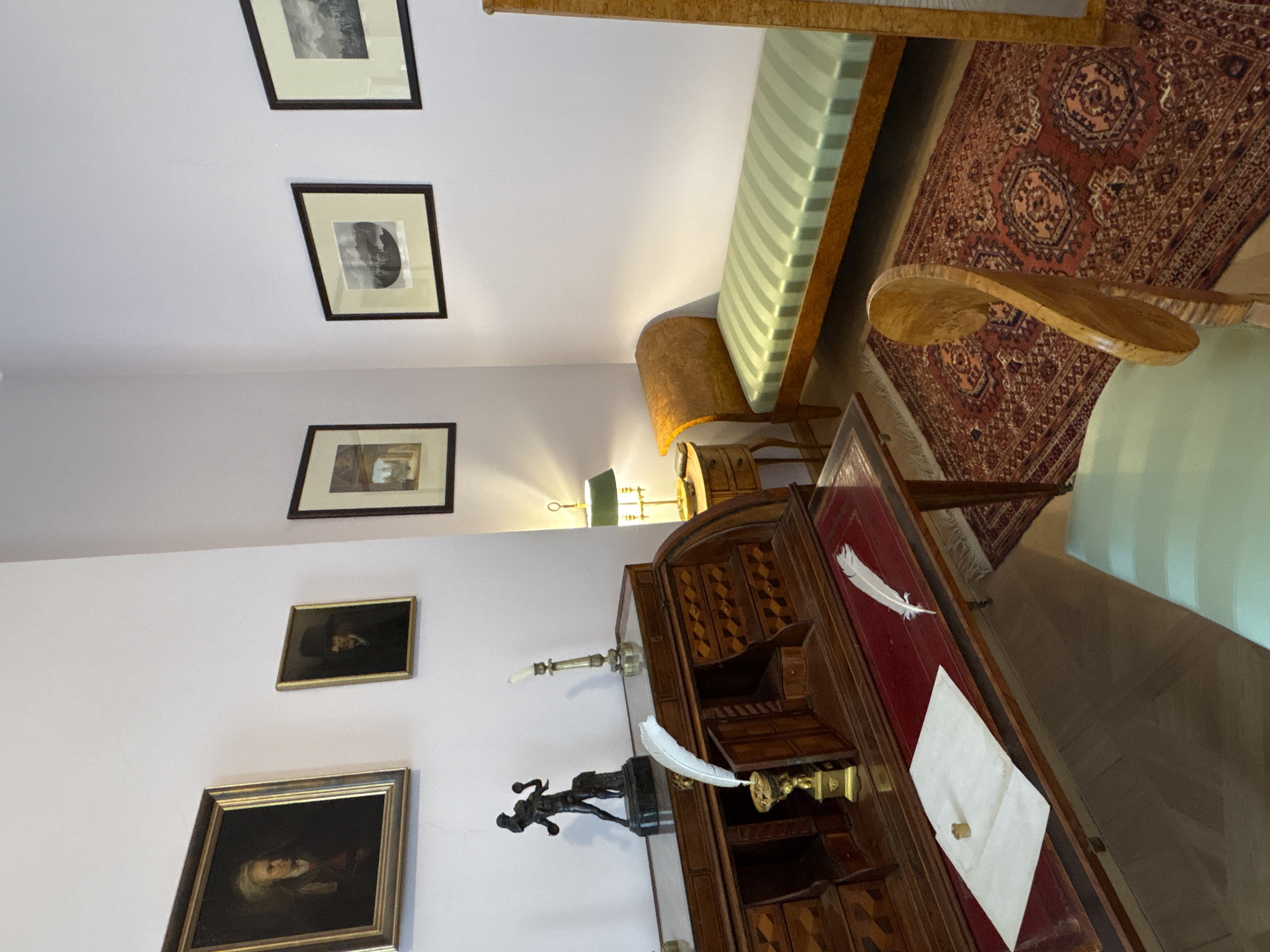 The image depicts a dimly lit room, possibly a study or a living area, furnished with antique pieces. On the left, a wooden roll-top desk with intricate inlays is prominent. It holds various items including a bronze sculpture of a figure, a candlestick, and writing implements such as quills and inkwells. A framed portrait hangs above the desk. To the right of the desk, a small bedside table supports a green-shaded lamp. Further to the right, a light-colored chaise lounge with green striped upholstery is positioned. The floor is covered with a large, intricately patterned oriental rug. In the foreground, a wooden chair with a light green seat is partially visible. The walls are painted a light color, and several framed artworks are displayed. The scene suggests a historical setting, potentially within a museum or a preserved historical residence.
This room is located in the Gatchina Palace, a former imperial residence in the city of Gatchina, Russia, located southwest of Saint Petersburg. Specifically, this room is believed to be the study of Pavel I.