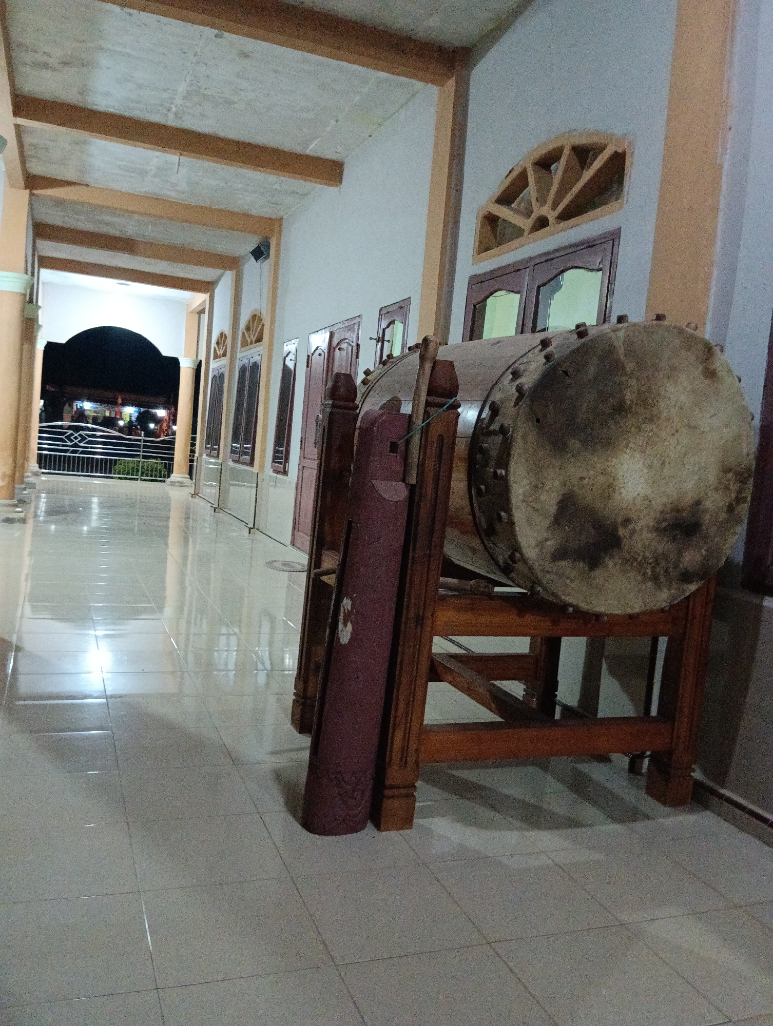 The image depicts a hallway or covered walkway of a building, likely a mosque or community center, in Pematang Jaya, Indonesia. The walkway has a tiled floor with a high sheen, reflecting the overhead lighting and the surrounding architecture. On the right side of the image, a large, traditional drum, possibly a bedug, is prominently displayed on a wooden stand. The drum has a weathered, skin-like surface secured by numerous studs around its circumference. Beside the drum, a rolled-up burgundy mat or banner with decorative patterns is leaning against a wooden support.
The hallway features columns supporting an overhead structure with exposed wooden beams. Along the left wall, a series of windows with arched decorative frames are visible, interspersed with doors. The far end of the hallway opens to an outdoor area, where a fence and some lights suggest a public space, perhaps a street or parking lot, with a car visible. The lighting in the hallway is artificial and casts long shadows, indicating it is likely evening or night. The overall impression is one of a quiet, well-maintained community space.