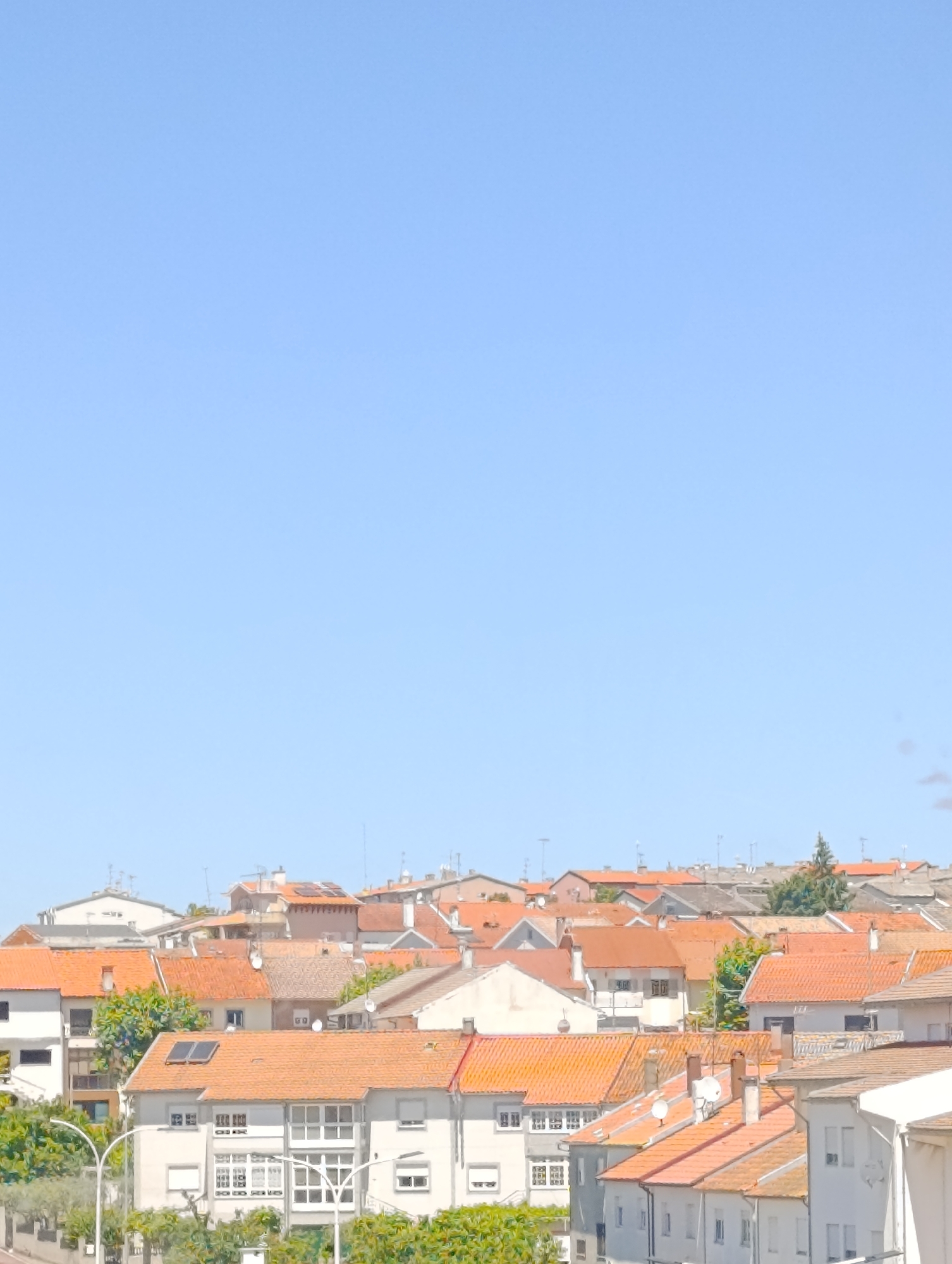 The image shows a cityscape under a clear blue sky. The location is Unknown, Unknown. The scene is dominated by buildings, mostly residential, with orange and grey tiled roofs. There are a few trees and some streetlights visible in the lower part of the frame. There are numerous TV antennas on the rooftops. The overall impression is that of a densely populated urban area.