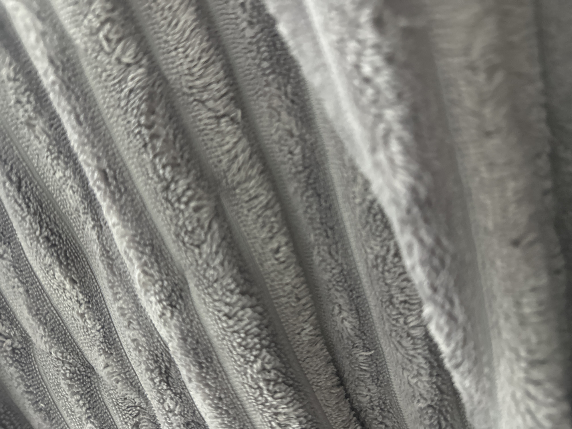 The image shows a close-up, abstract view of a grey, ribbed, fuzzy fabric. The fabric has a textured surface with raised lines running diagonally across the frame. The lighting creates subtle shadows and highlights, emphasizing the plush and soft appearance of the material. There are no people, actions, or specific locations visible in the image, making it impossible to determine its connection to Praskoveyevka, Russia.