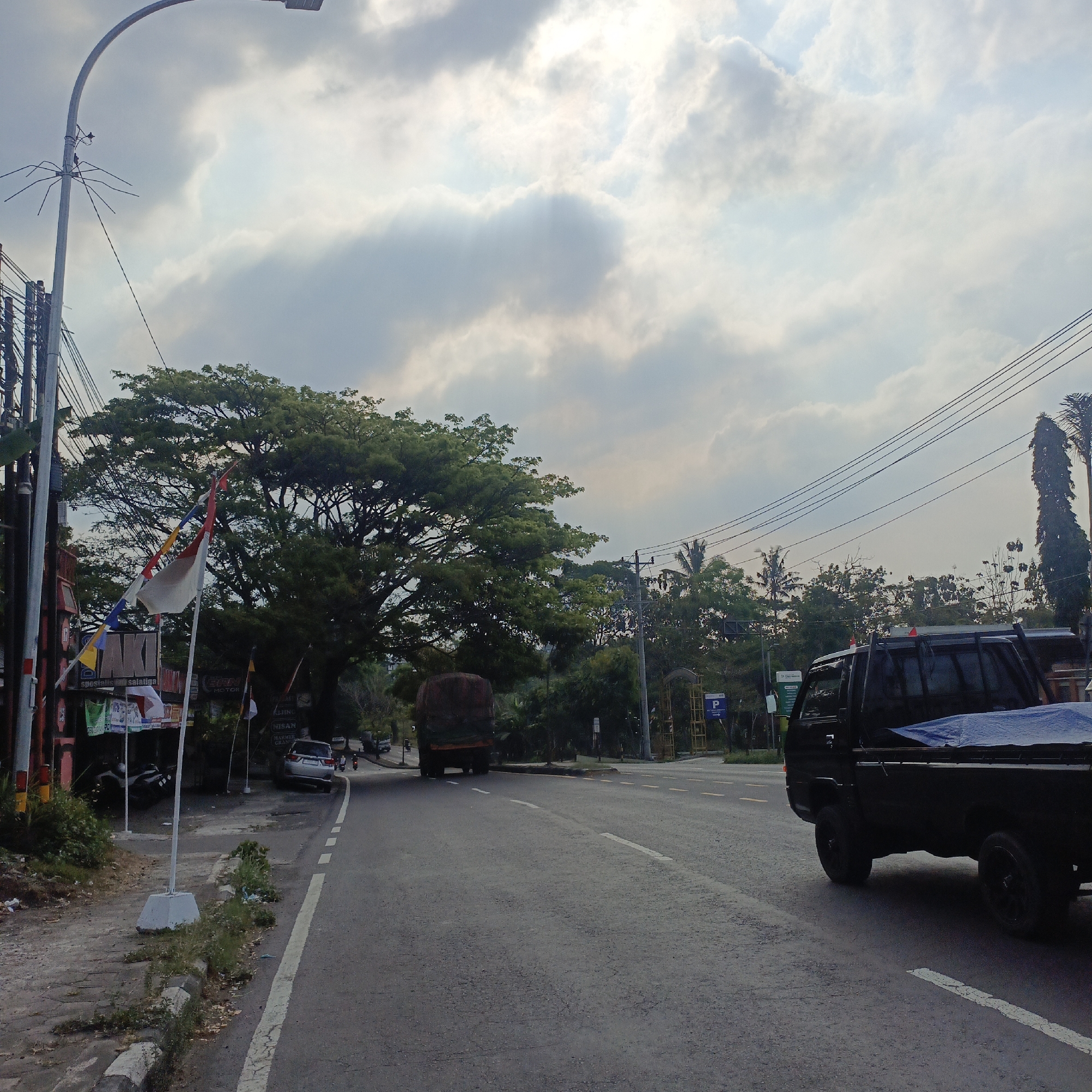 The image depicts a street scene in Salatiga, Indonesia, under a cloudy sky with rays of sunlight breaking through. A black pickup truck with a blue tarp covering its bed is parked on the right side of the road, facing away from the viewer. To the left of the road, a large tree with dense green foliage stands prominently. In the foreground on the left, a paved walkway is visible, with a white pole and flags. Further down the road, a large truck with a dark brown load is in motion, heading away from the viewer. Several other vehicles, including a silver car, are also present on the road. The environment includes power lines, streetlights, and various buildings and signage along the roadside.