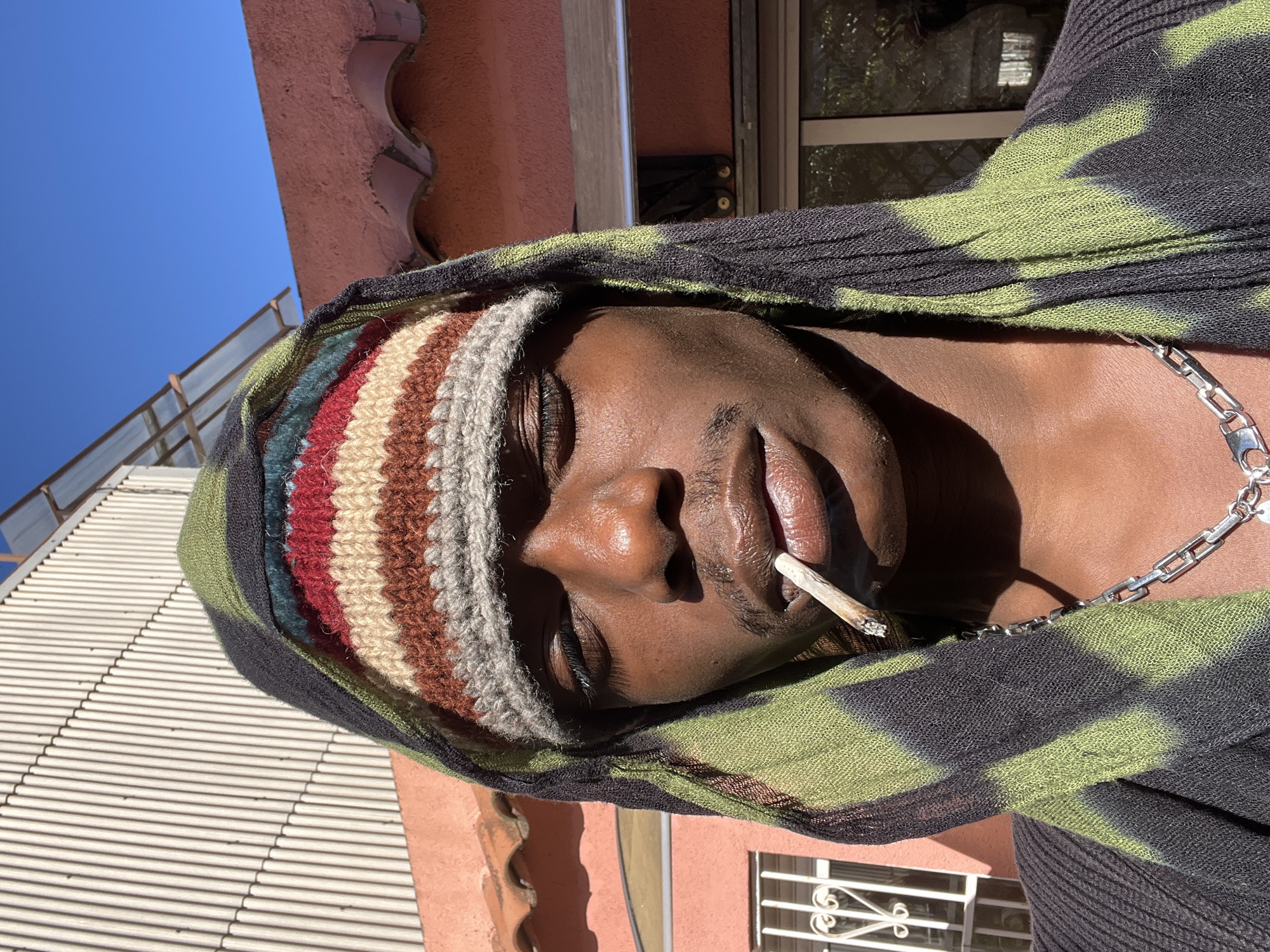 A man with dark skin, wearing a striped beanie and a patterned hood, is holding a lit cigarette in his mouth. He is also wearing a silver chain around his neck. The background includes a building with a terracotta-colored facade, a window with a wrought-iron grill, and a glimpse of a blue sky. The building and architectural details are consistent with architectural styles found in Barcelona, Spain.