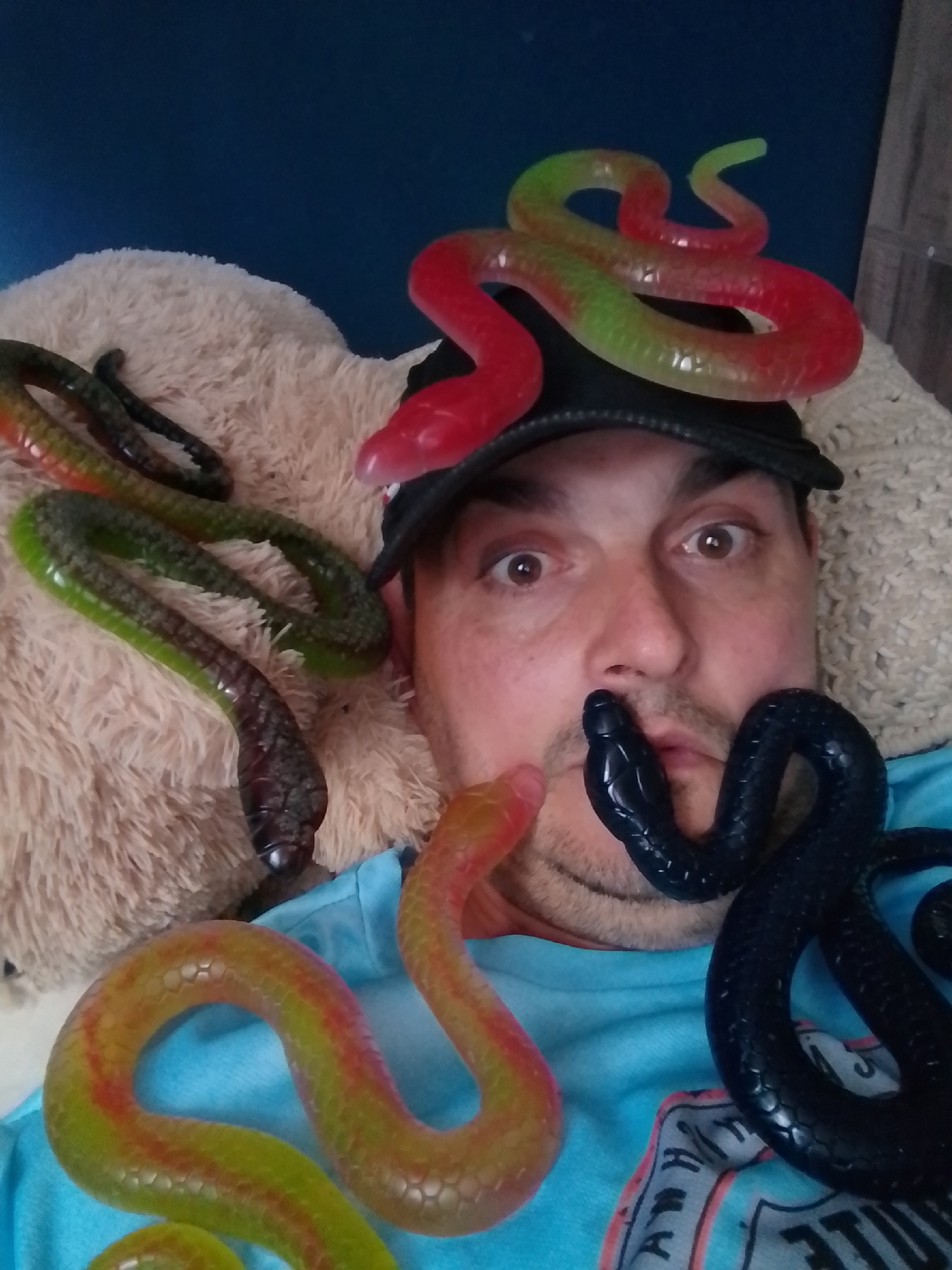 In an indoor setting with a blue background, a man wearing a blue t-shirt and a black baseball cap is the central subject of the image. He has brown eyes and stubble on his face. The man is surrounded by multiple colorful rubber snakes. One red and green snake is draped over his cap, another green and dark-colored snake lies to his left on a beige fluffy surface, a yellow and red snake is in the lower left of the frame, and a black snake is coiled near his face. The snakes appear to be toy replicas. The man's expression is neutral, and his eyes are wide open. The presence of plush items suggests a relaxed or playful environment. The image quality is moderate, with some graininess. There is no indication of the specific location within Wedel, Germany.