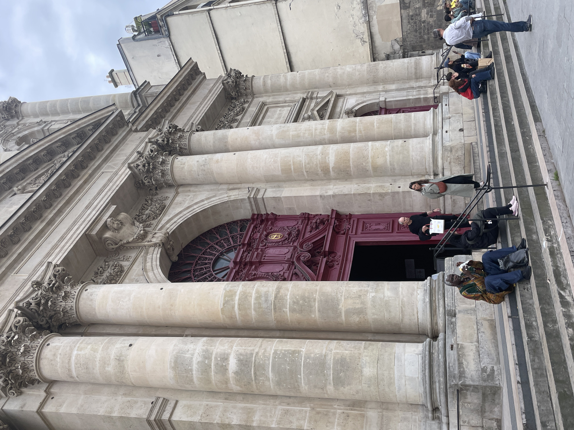The image depicts the entrance of a building, likely a church or a grand public building, in Paris, France. The architecture features large, ornate stone columns with Corinthian capitals and a prominent, dark red double door with intricate carvings and a decorative arched window above. In front of the entrance, a wide set of stone steps leads down to a paved area.
Several individuals are present in the scene. On the steps, a Black man wearing a colorful dashiki and blue jeans is seated, holding a piece of food and eating. To his right, a Black woman wearing a dark jacket is also seated. Further up the steps, a man in a black long-sleeved shirt and black pants is walking out of the building, carrying a white bag. A woman in a light-colored trench coat and dark pants is standing near the entrance.
Further down the steps, to the right, a group of people are seated. One woman with red hair and a red jacket is sitting with her legs crossed, and a person with a blue baseball cap is seated next to her. To their right, a man in a light blue shirt and jeans stands looking towards the right side of the frame. In the background, other people are visible seated or standing further away on the steps.
The environment appears to be an overcast day, with a cloudy sky visible in the upper portion of the image. Adjacent to the main building, a lighter-colored building with windows and balconies is visible. The overall impression is a bustling urban scene with architectural grandeur and people engaged in various activities on the steps of a significant Parisian landmark.