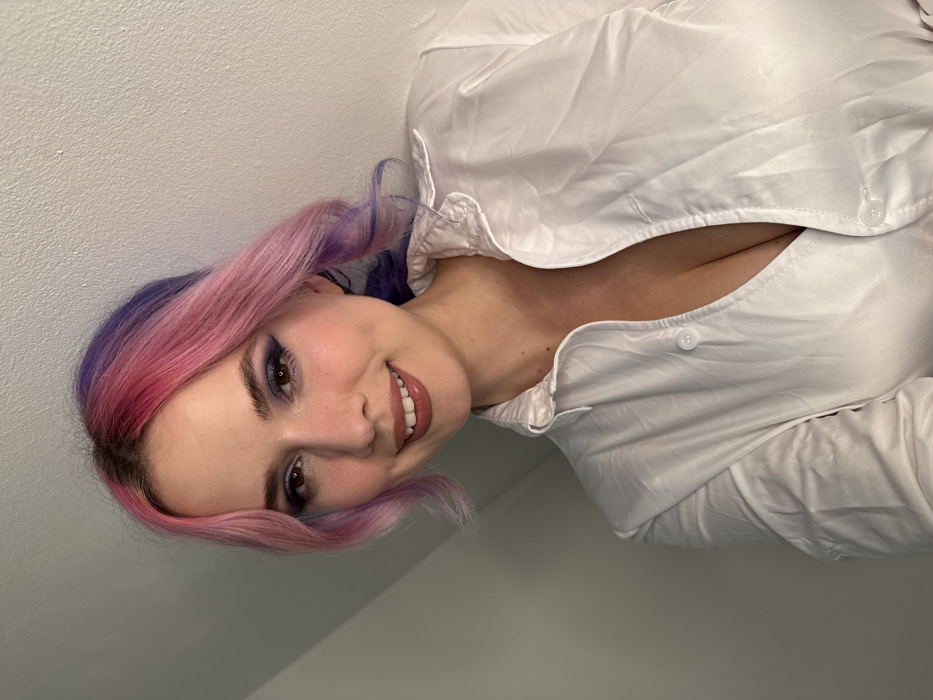 A close-up, waist-up indoor photograph depicts an adult female subject. The subject has multi-toned wavy hair, primarily pink near the scalp and transitioning to purple at the ends. Her face displays dark blue winged eyeliner, dark mascara, blush, and brownish-nude lip gloss. She is smiling, revealing teeth, and looking directly at the camera. She wears an unbuttoned white long-sleeved collared shirt, exposing cleavage. The background consists of a plain, off-white wall, with a diagonal ceiling line visible in the upper left quadrant. The specific location within the City of Zagreb, Croatia, is not discernible from the provided image.