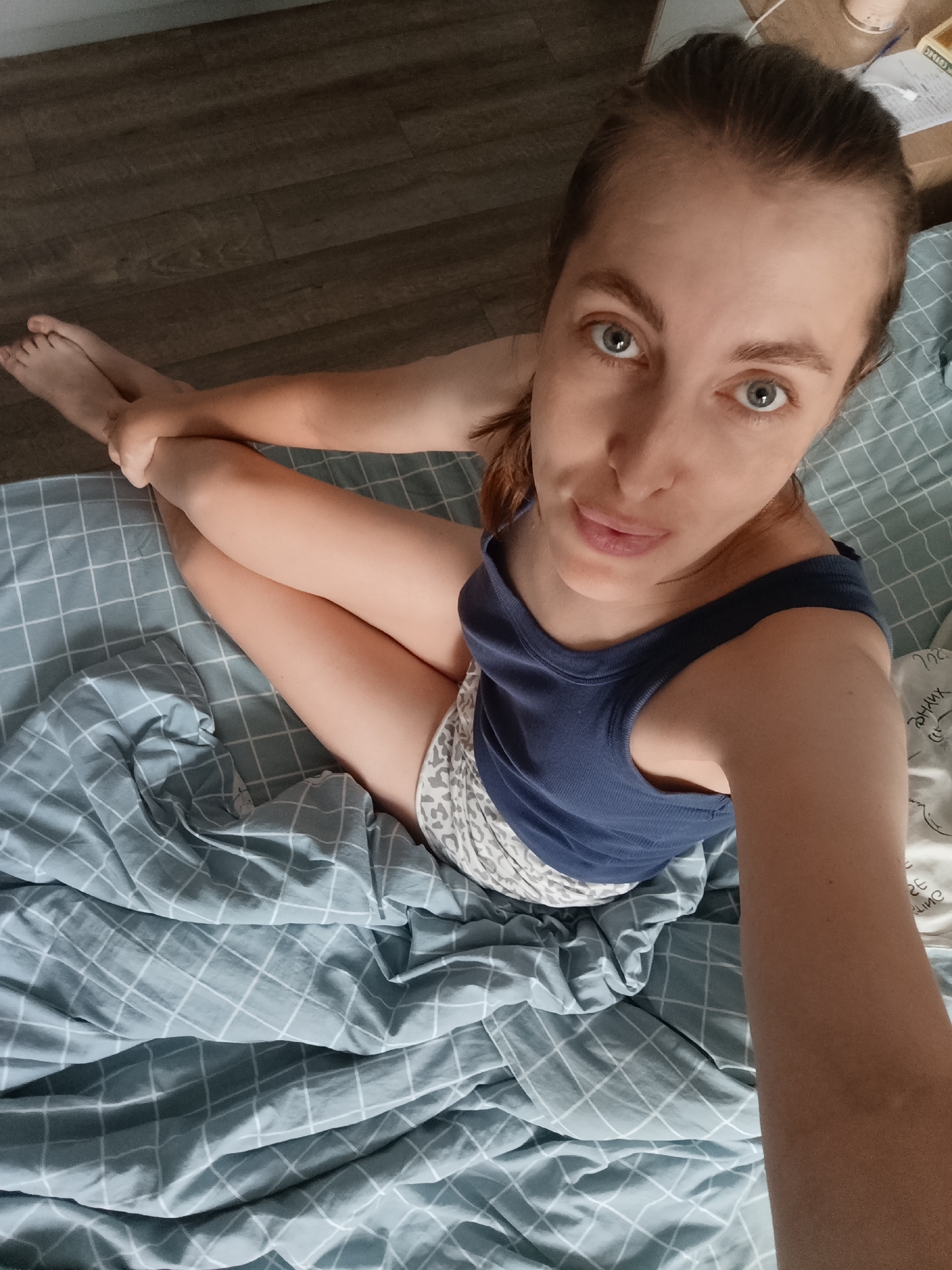 The image shows a woman sitting on a bed with her legs crossed. She is wearing a blue tank top and patterned shorts. Her hair is pulled back in a ponytail, and she has light blue eyes. The bed is covered with a light blue and white checkered comforter. The floor in the background appears to be dark wood. The image is a selfie, taken from a high angle. The location is not discernible from the image alone, and therefore cannot be identified as Chaikovsky, Russia.