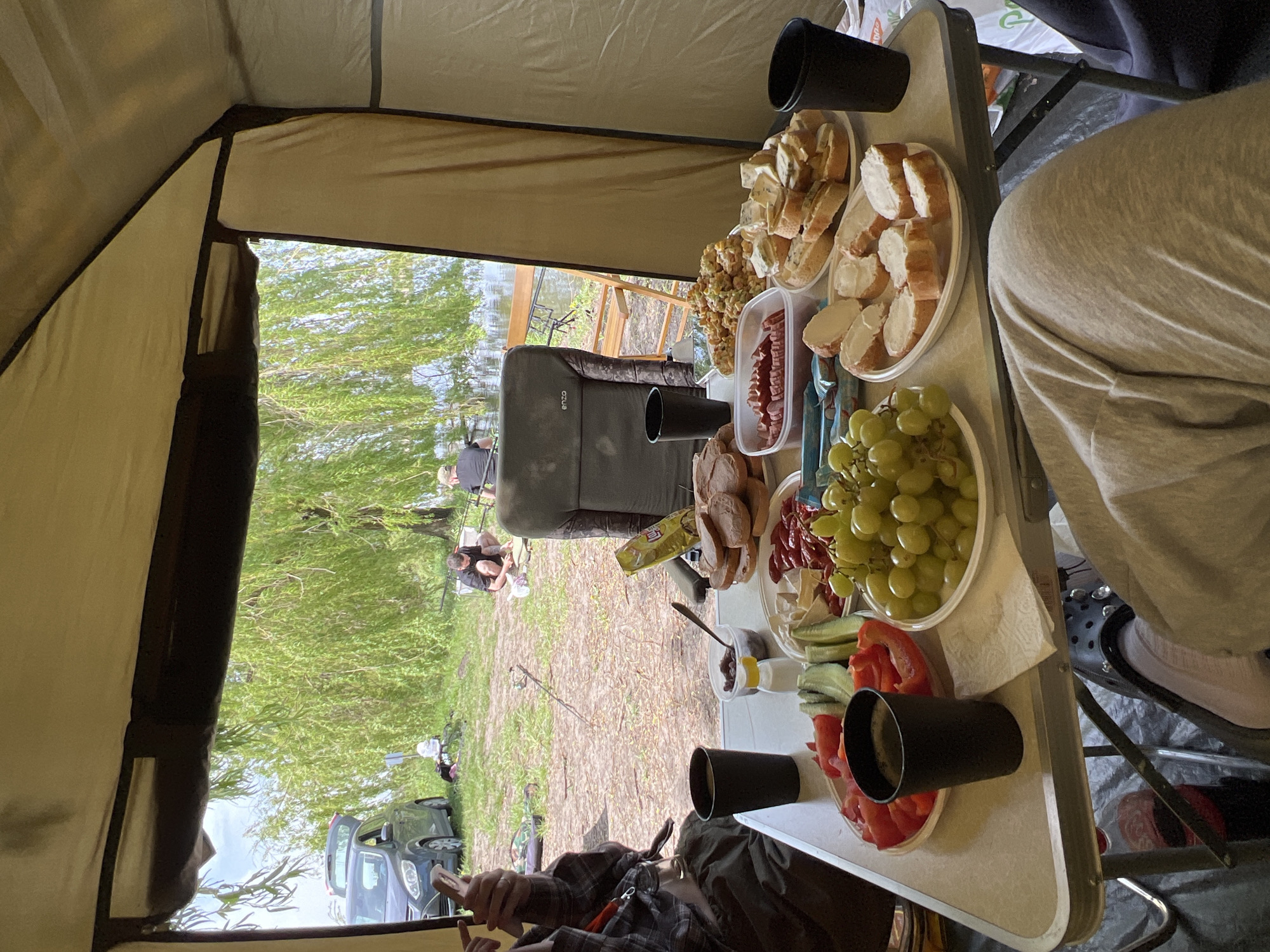 The image shows a view from inside a tent, possibly at a campsite near Romanove Selo, Ukraine. A folding table is set up inside the tent, laden with food, including sliced bread, grapes, sausage, peppers, and snacks. Black cups are also on the table. Outside the tent, the scene includes a grassy area, a body of water, trees, and at least two people who appear to be fishing. A gray vehicle is parked nearby. A fishing chair is visible near the center of the frame.