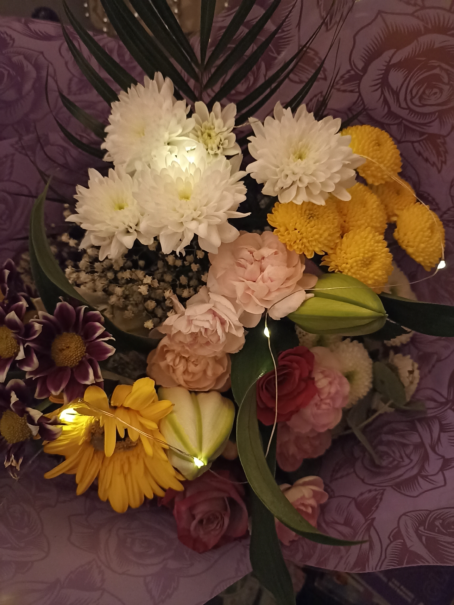 A bouquet of flowers is arranged on a purple floral patterned background. The bouquet contains white chrysanthemums, yellow mums, pink carnations, dark purple dahlias, yellow gerbera daisies, a red rose, and other smaller white and pink flowers. There are also some green leaves and a green decorative element that resembles a starfruit. Tiny LED string lights are woven through the bouquet. The overall lighting is dim, with the lights in the bouquet providing a warm glow. There are no people or animals present. The background appears to be wrapping paper or fabric with a repeating rose pattern. This scene is not specific to any location in Annagry, Ireland.