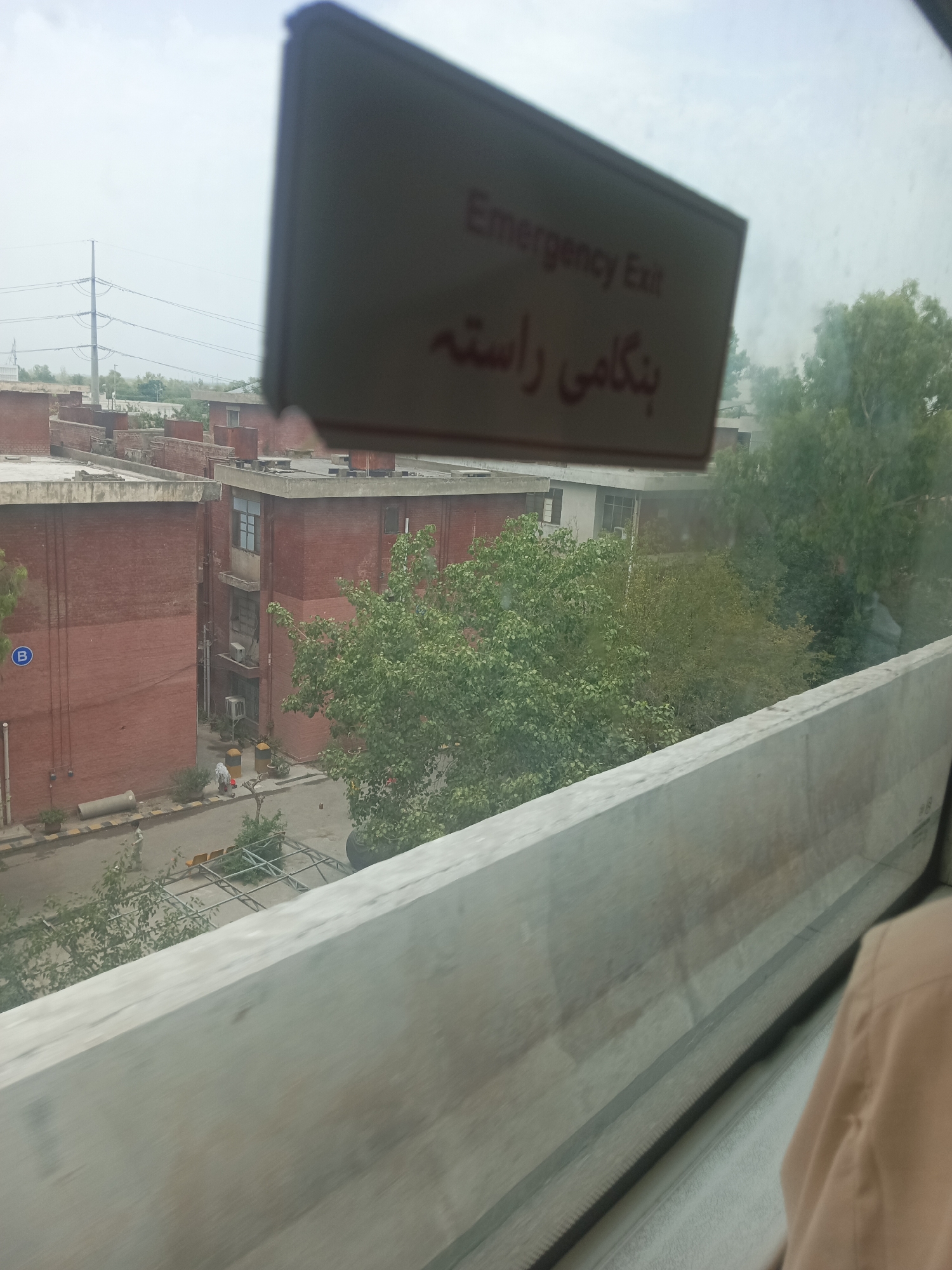 The image depicts an exterior view of brick buildings with multiple floors, likely an institutional or residential complex, under an overcast sky. In the foreground, a concrete barrier runs horizontally across the lower portion of the frame, obscuring the lower parts of the scene. A sign with the text "Emergency Exit" is visible, angled and partially obscuring the buildings behind it. The text "بنگامی راستہ" is written below "Emergency Exit" in what appears to be Urdu script. The buildings have various windows, some appearing to be air-conditioned units mounted on the exterior. Green trees are scattered around the buildings, with one particularly large tree to the right of the frame. A street or pathway is visible between the buildings, with some parked vehicles or equipment and a few individuals walking. The overall scene suggests a daytime view in a developed area, possibly a campus or housing society. The location provided is Alam Khan Kalay, Pakistan.