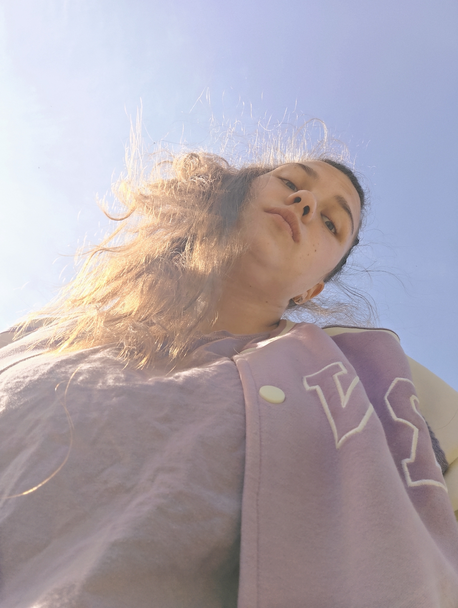 A low-angle, upward-facing photograph features a young woman against a clear blue sky. Her hair, a mix of brown and blonde, is somewhat disheveled and backlit by the sun. She is wearing a light purple top and a matching varsity-style jacket with white lettering and a single visible white button. The woman's face is slightly tilted, showcasing her nose and lips. The location is not possible to determine from this image.