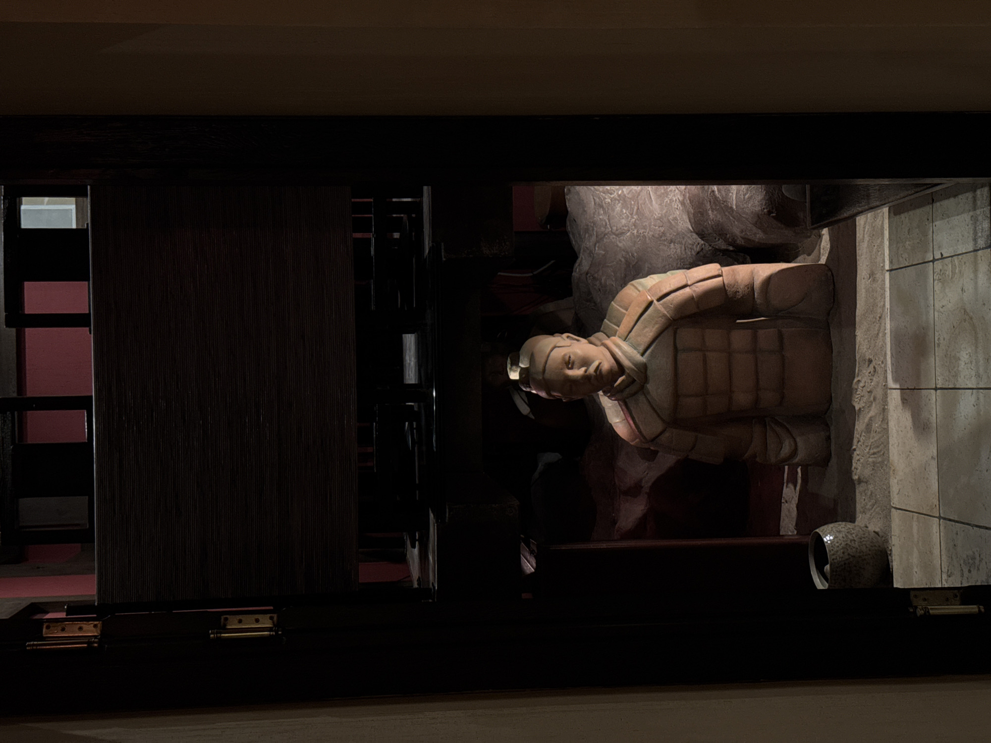 This vertically oriented image captures a static display of a terracotta warrior, likely in an exhibition or museum setting in Moscow, Russia. The view is framed by a dark, rectangular opening, resembling a window or part of a display case, suggesting the viewer is looking into an exhibit.
The central subject is a terracotta warrior statue, depicted from the chest upwards, with its head slightly turned towards the viewer. The warrior wears detailed, segmented armor in earthy tones of terracotta and beige. Its facial features are distinct, portraying a calm, stoic expression. The statue appears well-preserved, showcasing the intricate craftsmanship typical of the Terracotta Army.
Behind the warrior, the background is dimly lit, revealing textured, rock-like formations on the right side and a dark, possibly reddish, partition or wall on the left. A small, mottled ceramic pot or urn rests on the ground near the base of the display on the right. Further into the background on the left, a glimpse of another section of the exhibit can be seen, with a lighter-colored wall or object.
The overall scene is characterized by dramatic, low lighting, highlighting the warrior statue while keeping the surrounding elements largely in shadow. There is no visible activity, and no text is discernible within the frame. The image conveys a sense of quiet observation of a historical artifact.