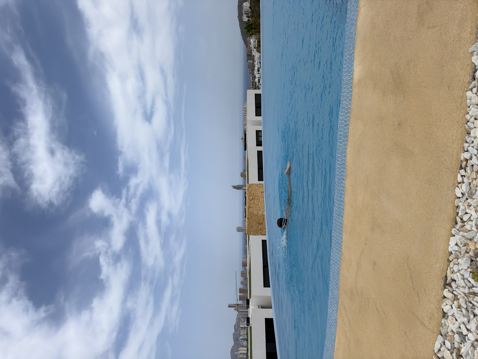 A person swims in an infinity pool on a rooftop in Finestrat, Spain. The person is wearing a swimming cap and goggles and is submerged in the blue water, with their head and shoulders visible. The pool has a tan colored edge with small white tiles. In the background, a city skyline with tall buildings and mountains can be seen under a cloudy blue sky. The scene captures a moment of relaxation and leisure in an urban coastal environment.