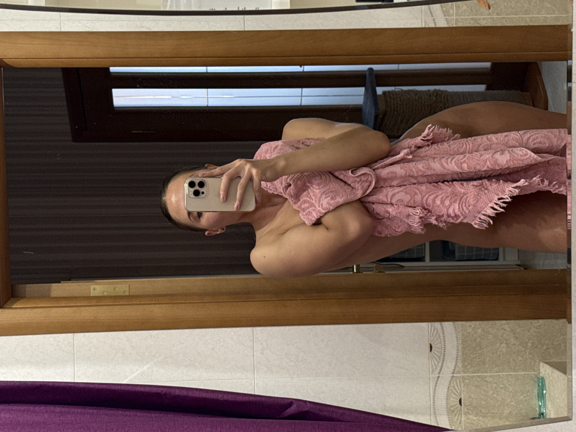 A person is captured in a mirror selfie within what appears to be a bathroom setting. The individual, seemingly female, is wrapped in a textured, light pink towel adorned with a subtle damask pattern. The towel covers her torso and legs, leaving her shoulders and arms exposed. She holds a light-colored smartphone, likely an iPhone with a triple-lens camera, in her right hand, which obscures the lower part of her face. Only her left eye, forehead, and part of her cheek are visible, showing a neutral expression.
The reflection reveals a room with tiled walls; some sections are plain white, while others feature decorative beige tiles with a repeating fan-like motif. A wooden frame, possibly part of the mirror's border or a piece of furniture, is prominent in the foreground. In the background of the reflection, a window with horizontal blinds or slats is visible, letting in some ambient light. There's also a dark, possibly wooden, surface and a textured brown object. A purple fabric or curtain is visible at the bottom of the mirrored frame, suggesting it's part of the room's decor. The image is taken with indoor lighting, and no specific time of day or external weather conditions can be discerned. There is no visible text.