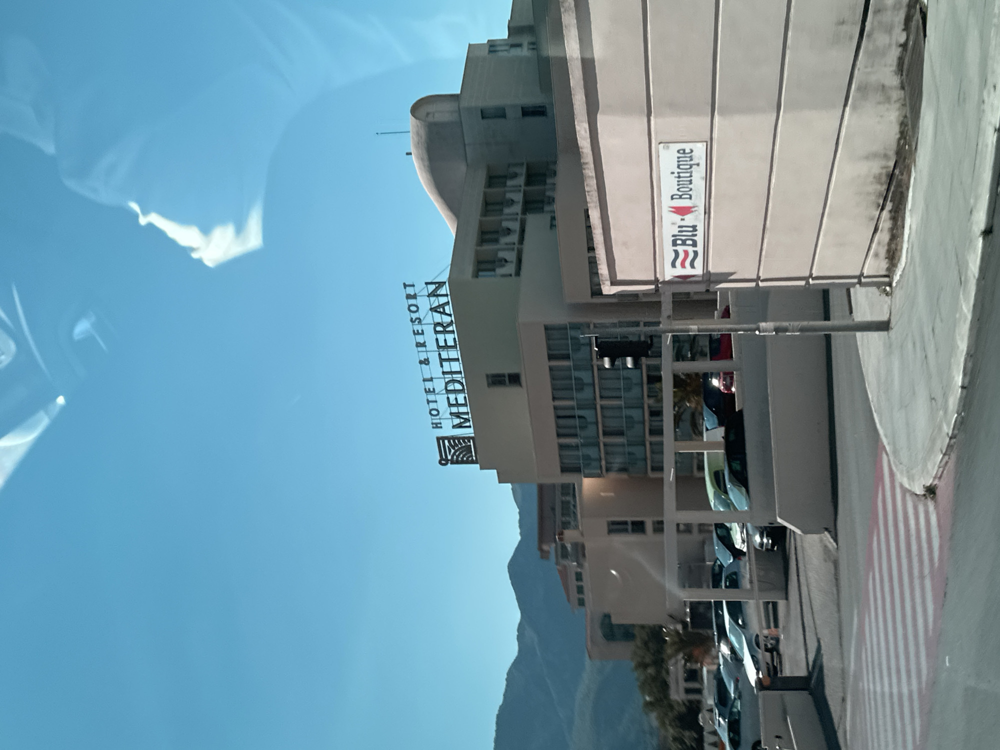 The image captures a sunny day in Budva, Montenegro, with a clear blue sky and distant mountains. The main subject is the "HOTEL & RESORT MEDITERAN," a large building complex with a distinctive curved roof. The hotel sign is prominently displayed on the top of the building. In the foreground, a parking lot is visible, filled with various cars. A traffic light stands at an intersection, and a pedestrian crossing with red and white stripes is present. To the right of the hotel, a wall features a sign for "Blu Boutique." The overall scene depicts a typical urban environment with commercial establishments and parking facilities.