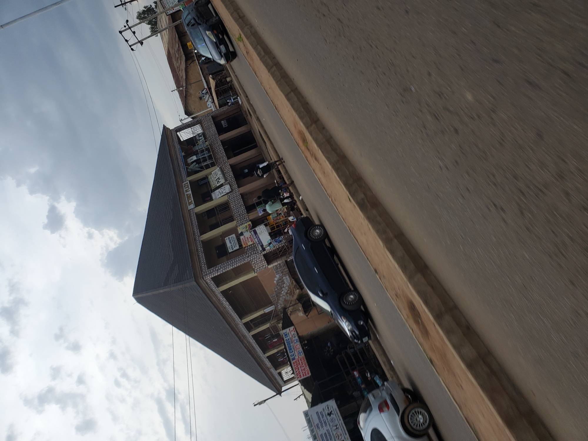 This image captures a street scene in Bukuru, Nigeria, on a cloudy day. In the foreground, a paved road runs diagonally across the frame, with a raised concrete curb separating it from a dirt shoulder. To the left, a silver car is parked on the shoulder, partially obscured. The main subject of the image is a multi-story building with a dark, sloped roof. The building has a light-colored facade with decorative stonework on the lower levels and balconies on the upper level, where several people are visible standing and interacting. Various signs are affixed to the building, including "VICTORY PLAZA," "MEDICAL LABORATORY," and others that are difficult to read. Further down the street, a black car is driving away from the camera. The background shows more buildings with corrugated metal roofs and utility poles with overhead wires. The sky is overcast with dramatic cloud formations. The overall impression is of a bustling urban environment.