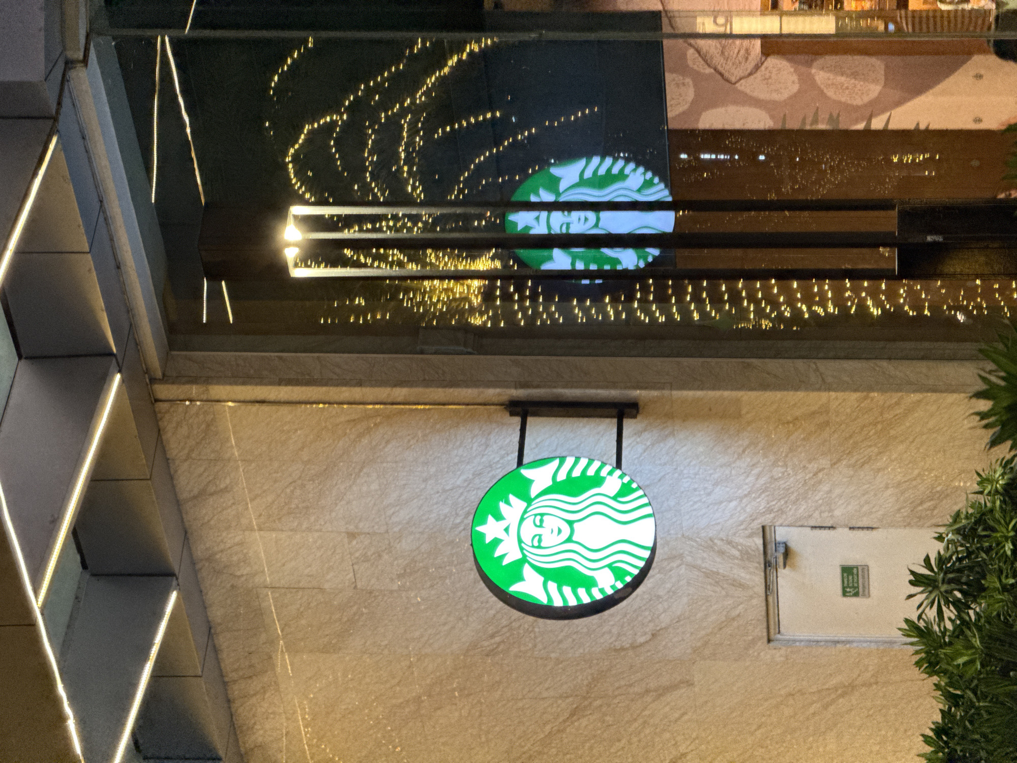 The image captures a Starbucks signage outside a building at night. The circular green and white Starbucks logo is illuminated and mounted on the building's facade. To the right, the logo is reflected in a large glass window, behind which a string of warm-toned fairy lights is visible, creating a festive ambiance. The building exterior is clad in textured beige stone. In the lower left, lush green foliage is present. Below the Starbucks sign, a plain white door with a green emergency exit sign is visible. The scene appears to be set in an urban environment, possibly a commercial district. The context of Nha Trang, Vietnam, suggests a location within this coastal city.