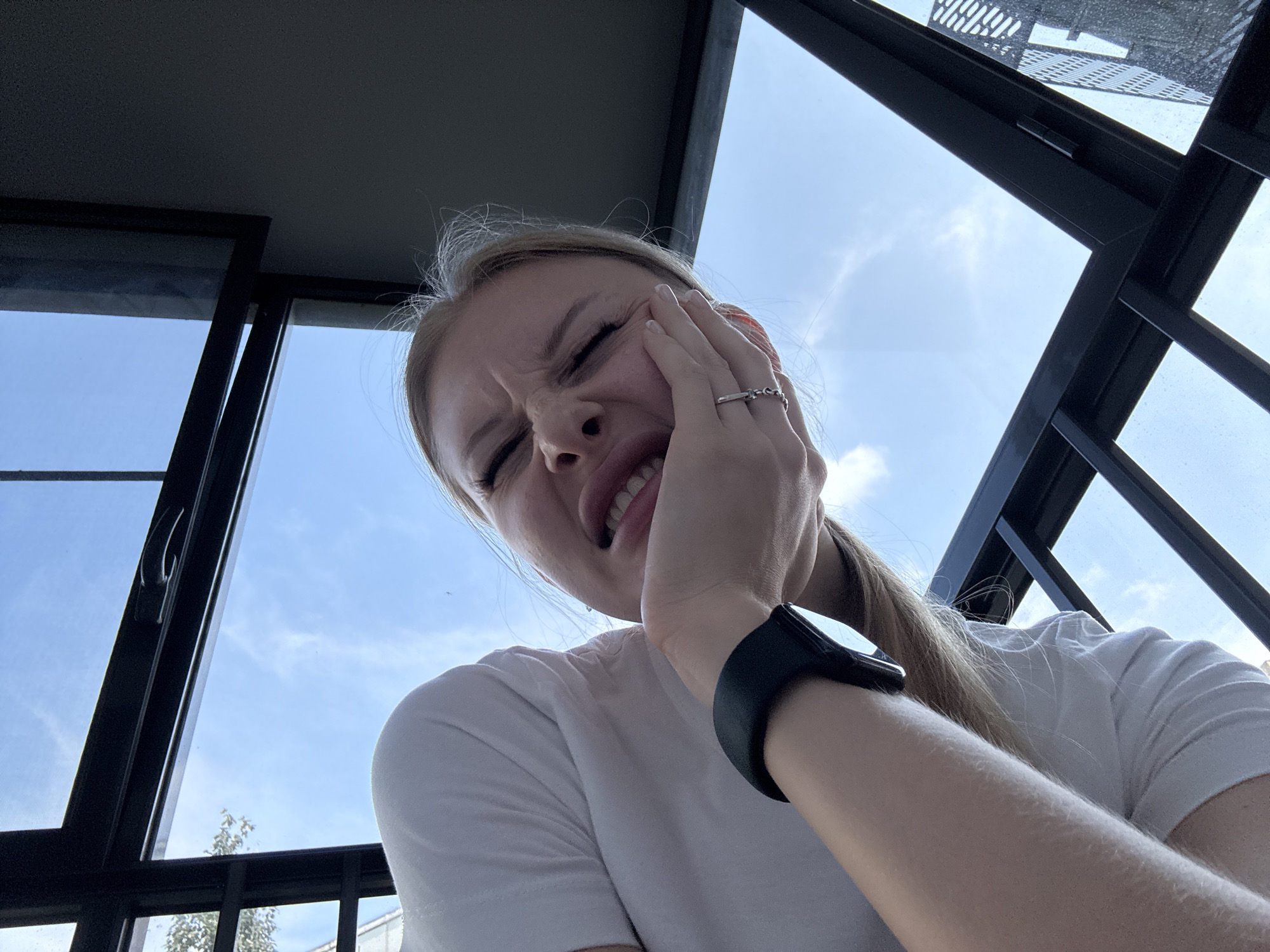 A young Caucasian woman is captured from a low angle, looking upwards. She is wearing a white t-shirt and a black wristwatch. Her right hand is pressed against her cheek, and her eyes are closed with a grimace on her face, suggesting discomfort or pain. Her mouth is partially open, revealing her teeth. She has long blonde hair. The background consists of a blue sky with scattered clouds, framed by dark metal structures, possibly part of an outdoor seating area or building exterior. The overall impression is that of a candid moment, possibly taken outdoors on a sunny day. There is no information in the image to suggest the location is the city of Perm, Russia.