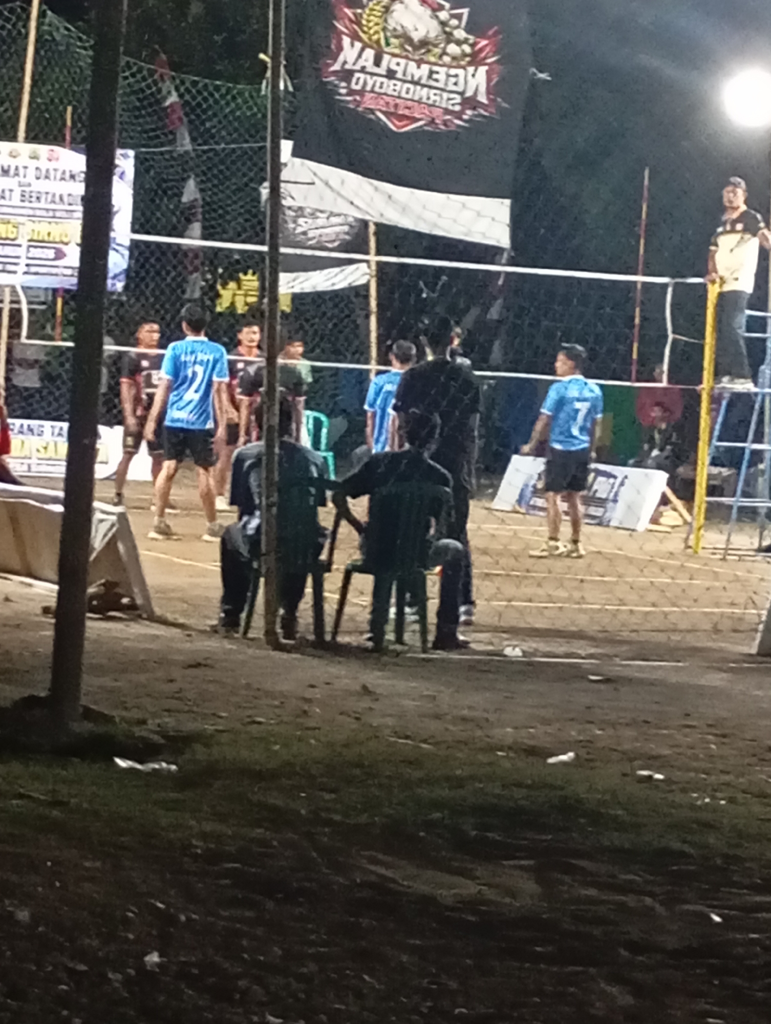 The image shows a group of young men on a volleyball court at night. They are wearing athletic clothing, and some are holding volleyballs. The court is lit by artificial lights, and there is a net in the middle of the court. In the background, there is a banner with writing on it. The image appears to have been taken in a public area, possibly a park or a sports complex, in the city of Pacitan, Indonesia. The atmosphere is casual and energetic, with the players seemingly engaged in a game or practice session.