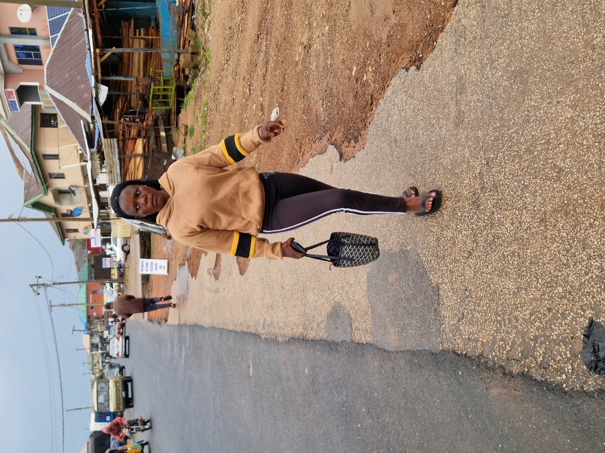 A woman walks on a paved sidewalk next to a road in Kabong, Nigeria. The woman is wearing a tan hooded sweatshirt with black and yellow stripes on the sleeves and black leggings with white stripes. She is carrying a black and beige patterned handbag in her right hand. Her feet are bare, and she is wearing black flip-flops. The sidewalk is wet in patches, indicating recent rain. On the other side of the road, there are shops and buildings, some with corrugated metal roofs. Vehicles, including a van and motorcycles, are visible on the road. The sky is overcast, suggesting cloudy weather. The ground on the side of the road is a mix of dirt and gravel.