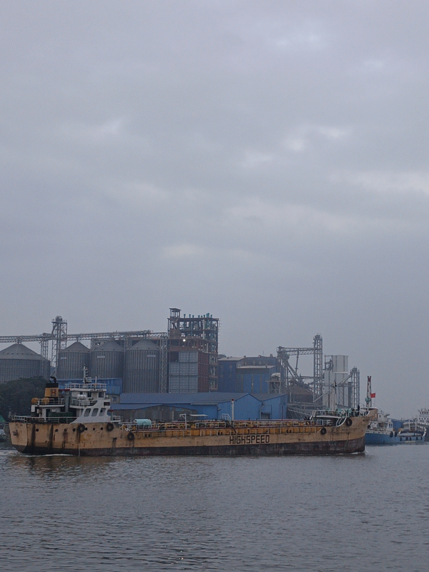 The image shows a hazy, overcast day at a port in Dhaka, Bangladesh. A large, tan tanker ship named "HIGHSPEED" dominates the midground, moving across the water. Behind the ship, a complex industrial area is visible, featuring grain silos, metal structures, and blue buildings. In the background, other ships are partially visible. The water appears calm and gray.