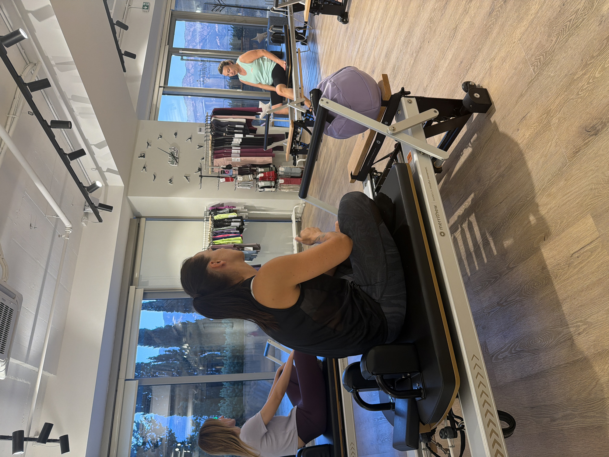 The image depicts the interior of a brightly lit Pilates studio, likely located in Boreti, Montenegro, given the context of the user's prompt. The studio features large windows offering a view of a landscape with trees and hills, suggesting a scenic setting.
In the foreground, a woman with dark hair pulled into a ponytail is seated on a Pilates reformer. She is wearing a black sleeveless top and grey patterned leggings, and appears to be in the middle of an exercise. To her left, another woman with blonde hair is also seated on a Pilates reformer, facing away from the camera. She is wearing a light purple t-shirt and dark purple leggings. Both women are positioned on their respective reformers, engaged in what appears to be a Pilates class or session.
Further back in the studio, a third woman, wearing a light blue tank top and black shorts, is seated on a Pilates reformer, looking towards the left of the frame. Behind the women, the studio is equipped with multiple Pilates reformers. Racks of Pilates apparel, such as leggings and tops in various colors, are visible on display. The floor is a light-colored wood, and the ceiling has a modern lighting system with track lights. A large, round, lavender-colored ball is resting on the side of one of the reformers. The overall environment suggests a professional and well-equipped fitness facility.