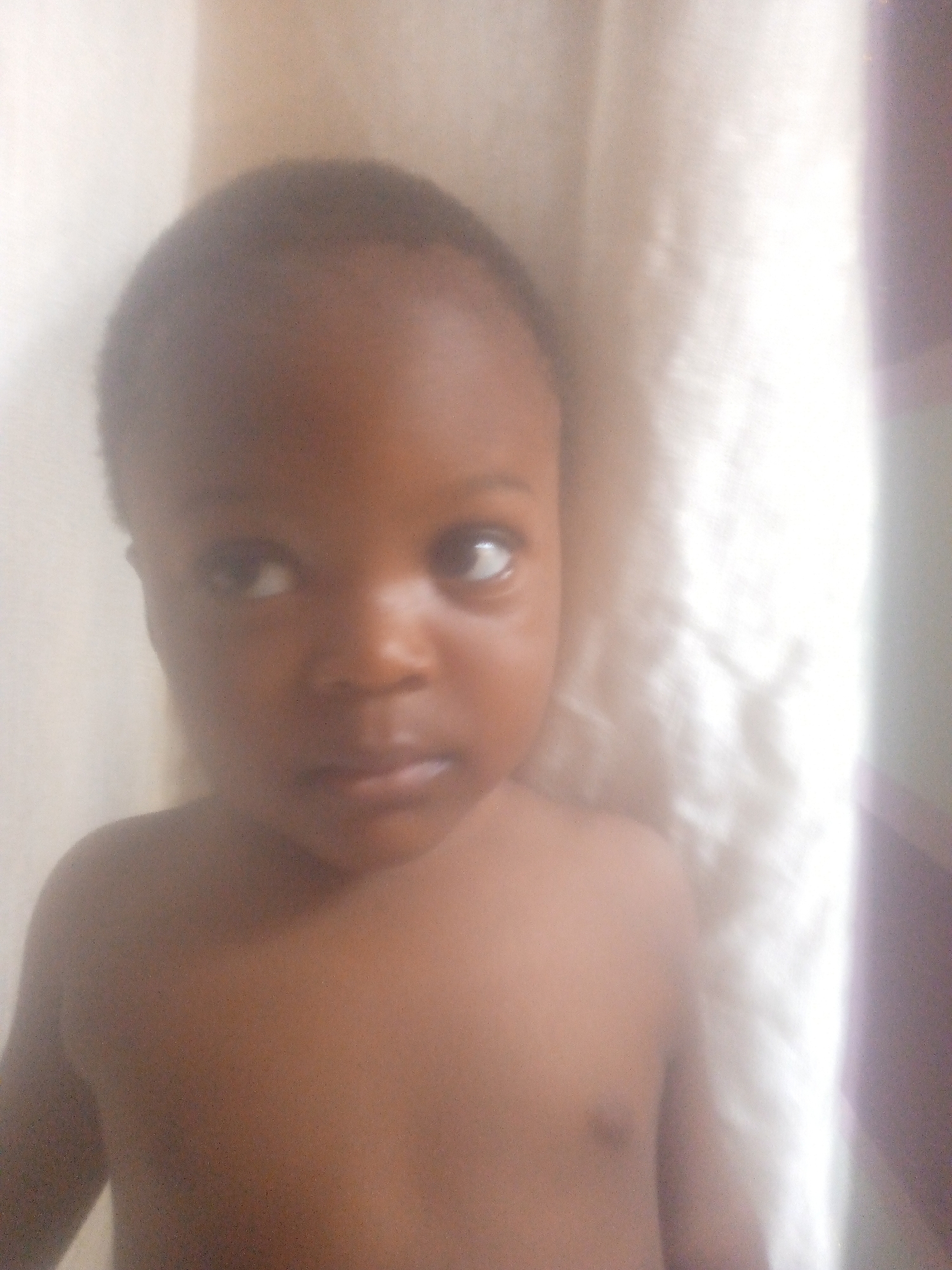 A close-up, indoor shot focuses on a young, dark-skinned child, appearing shirtless. The child's gaze is directed to the left, away from the camera. Behind the child is a textured white backdrop that could be fabric or a wall. The location of the scene is listed as Unknown, Unknown.