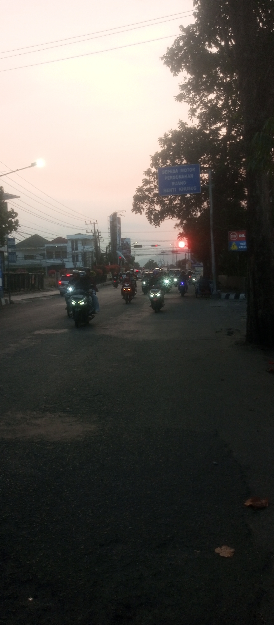 The image depicts a street scene in Kediri City, Indonesia, at what appears to be dusk or dawn. A road with multiple lanes is visible, with several motorcycles driving in the same direction. The motorcycles have their headlights on, illuminating the road. Buildings line the left side of the street, and a large tree with dark foliage is on the right. A blue sign with white text, possibly a traffic sign, is mounted on a pole. The sky is a light, hazy color, suggesting the sun is low on the horizon, and the sun itself is visible as a bright, red orb in the distance, slightly above the treeline. Traffic lights and streetlights are also present. The road surface appears to be asphalt and shows signs of wear.