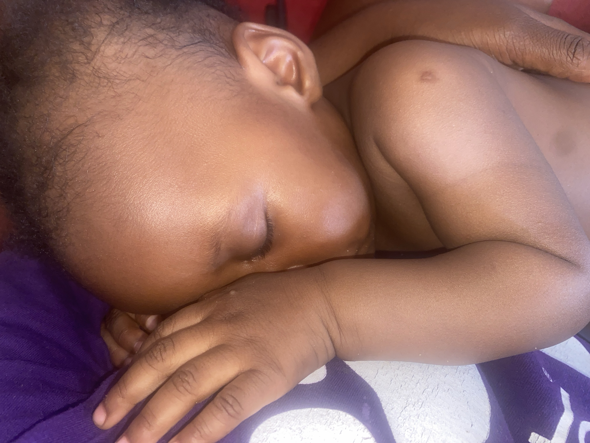 The image captures a close-up view of a young child, likely a baby or toddler, sleeping peacefully. The child has rich, dark skin and fine, dark hair. They are lying on their right side, with their face partially obscured by their right hand which is resting near their chin. Their eyes are closed, indicating sleep.
A larger, adult hand with visible wrinkles is gently placed or resting behind the child's upper back or shoulder, suggesting comfort, support, or a protective embrace. The child's left arm is also visible, resting on a surface. On the child's right upper arm or shoulder area, there is a small, faint, reddish-brown mark or mole.
The child is resting on a surface covered by two distinct fabrics: a vibrant purple fabric is visible under the child's chin and right hand, and a lighter, possibly white or off-white, textured fabric is beneath their left arm and extends further into the background. Small, light-colored speckles, possibly dust or lint, are visible on the child's skin and the fabrics in various parts of the image.
The lighting in the image is soft and warm, highlighting the child's smooth skin and creating a serene and intimate atmosphere. The overall impression is one of tranquility and tender care. There is no visible text in the image.