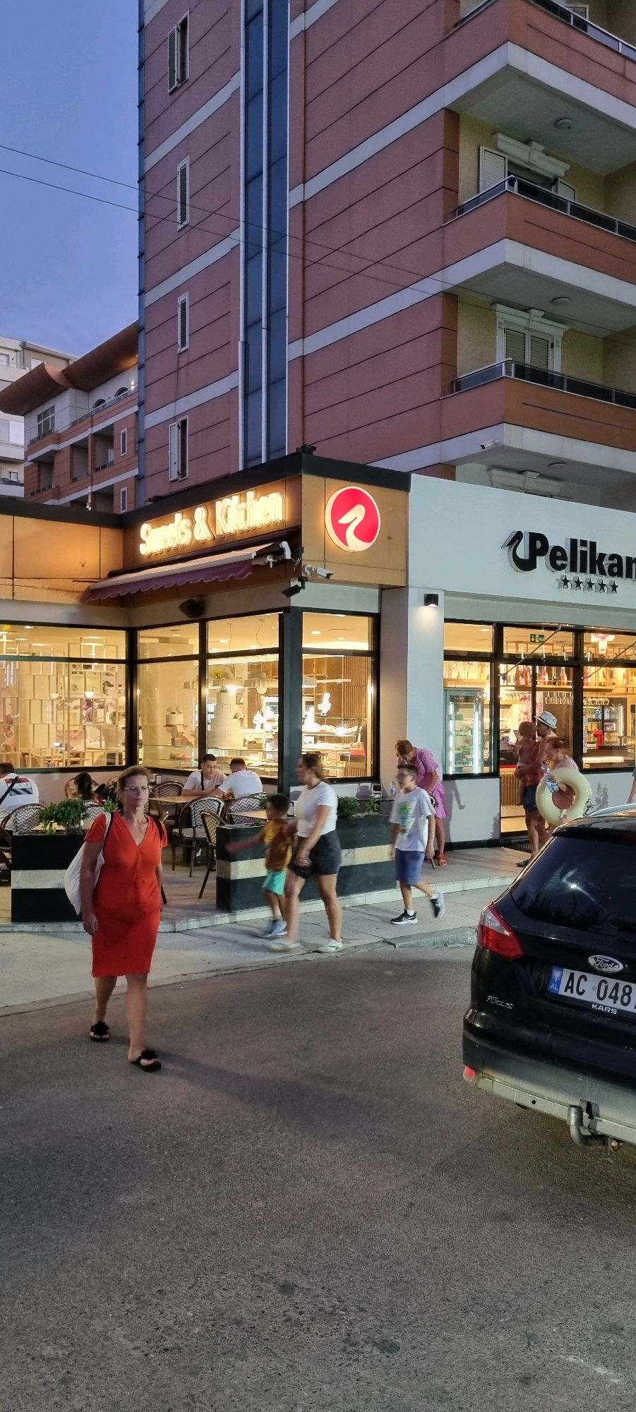 The image captures a street scene in front of a commercial establishment in Bashkia Durrës, Albania, likely during the late afternoon or early evening, judging by the lighting and the presence of illuminated signage. The dominant structure is a modern building with multiple stories, featuring balconies and a facade composed of pinkish-red panels interspersed with white horizontal bands and a central vertical blue glass section.
At the ground level, a commercial space with large glass windows is visible. A prominent sign reads "Sweets & Kitchen" in illuminated golden lettering. Adjacent to this is another establishment with a sign displaying "Pelikan" along with five star symbols. The interior of these businesses appears to be well-lit and stocked with items, suggesting they are shops or cafes.
Several individuals are present on the sidewalk and street. In the foreground, a woman in an orange dress walks towards the left of the frame, carrying a white bag over her shoulder. Behind her, a group of people, including children, are walking towards the right. One child, wearing a white t-shirt and blue shorts, walks with an adult female in a dark skirt and white top. Another child, in a yellow shirt and green shorts, is with them. Further to the right, a man in a hat holds a child and a yellow inflatable ring. In the background, several people are seated at tables outside the "Sweets & Kitchen" establishment.
A black Ford Focus car is parked on the right side of the street, facing away from the viewer. Its rear license plate is visible, showing "AC 048" followed by what appears to be an Albanian registration code "KARS". The street surface is asphalt. The overall environment suggests a commercial district with pedestrian activity.