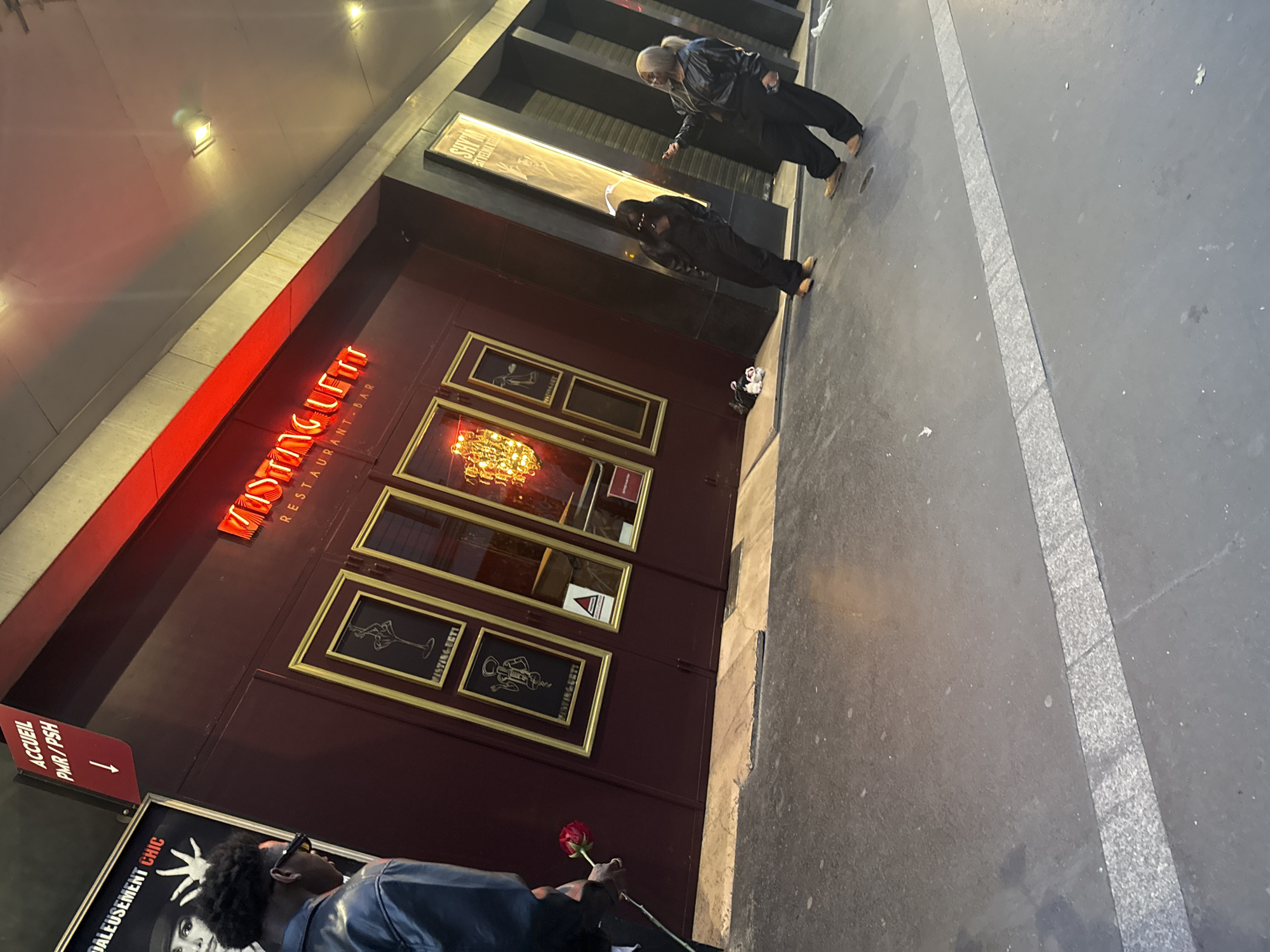 The image shows the exterior of a restaurant named "Mistinguett Restaurant-Bar." It is presumably located in Paris, France, considering the French words "ACCUEIL PMR/PSH" on a red arrow sign, meaning "Accessibility for People with Reduced Mobility/People with Disabilities." There are four framed pictures hanging on the wall, beneath the restaurant's name in red neon lights. A man holds a red rose in the lower part of the image. Further up, two people are standing or walking on the pavement adjacent to the building. A sign with the name "SHYM" can also be seen above the entrance. The sidewalk pavement is visible with a light gray stripe dividing the sidewalk.