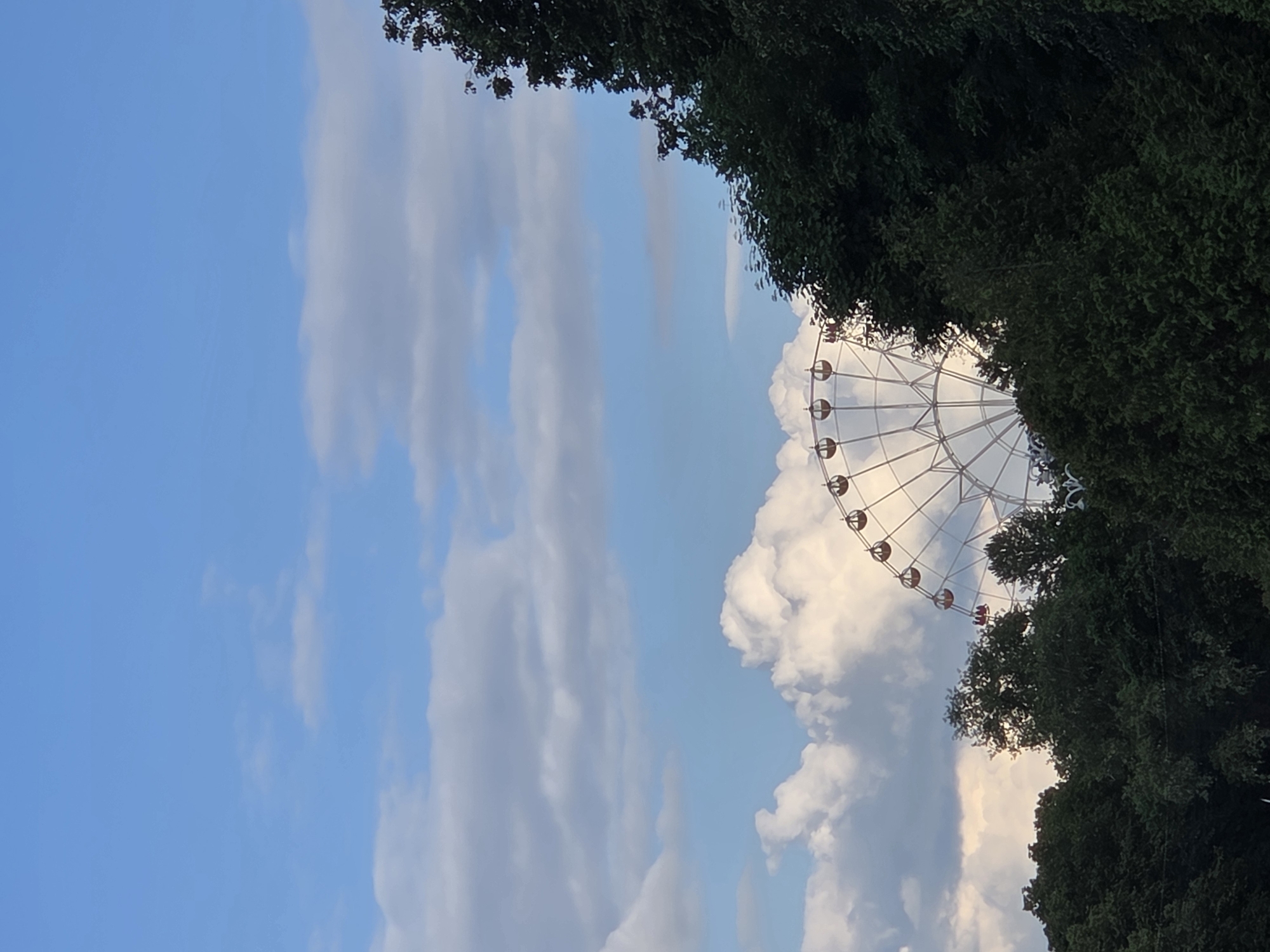 The image captures a Ferris wheel partially obscured by trees and clouds. The sky is a bright blue with scattered white clouds. The Ferris wheel is large and white, with several gondolas visible. It appears to be located in an outdoor setting, possibly a park or amusement area, amidst lush green trees. The overall impression is one of a serene, possibly late afternoon or early evening scene. It is impossible to determine the specific location in Minsk, Belarus from the image alone.