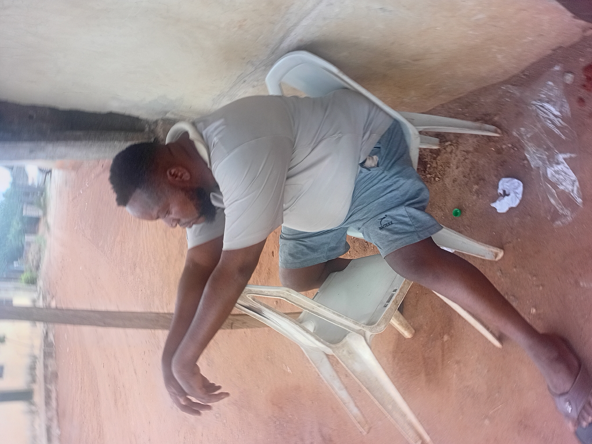 A man is depicted slumped over two white plastic chairs on a dirt surface in an outdoor environment. The man is wearing a light-colored t-shirt and grey shorts. He is dark-skinned with a beard and short hair. His arms are hanging down. The background consists of a light-colored wall, possibly a building exterior. A green round object and a piece of white cloth or paper are visible on the ground near the chairs. Based on the prompt, the location is Ikorodu, Nigeria.