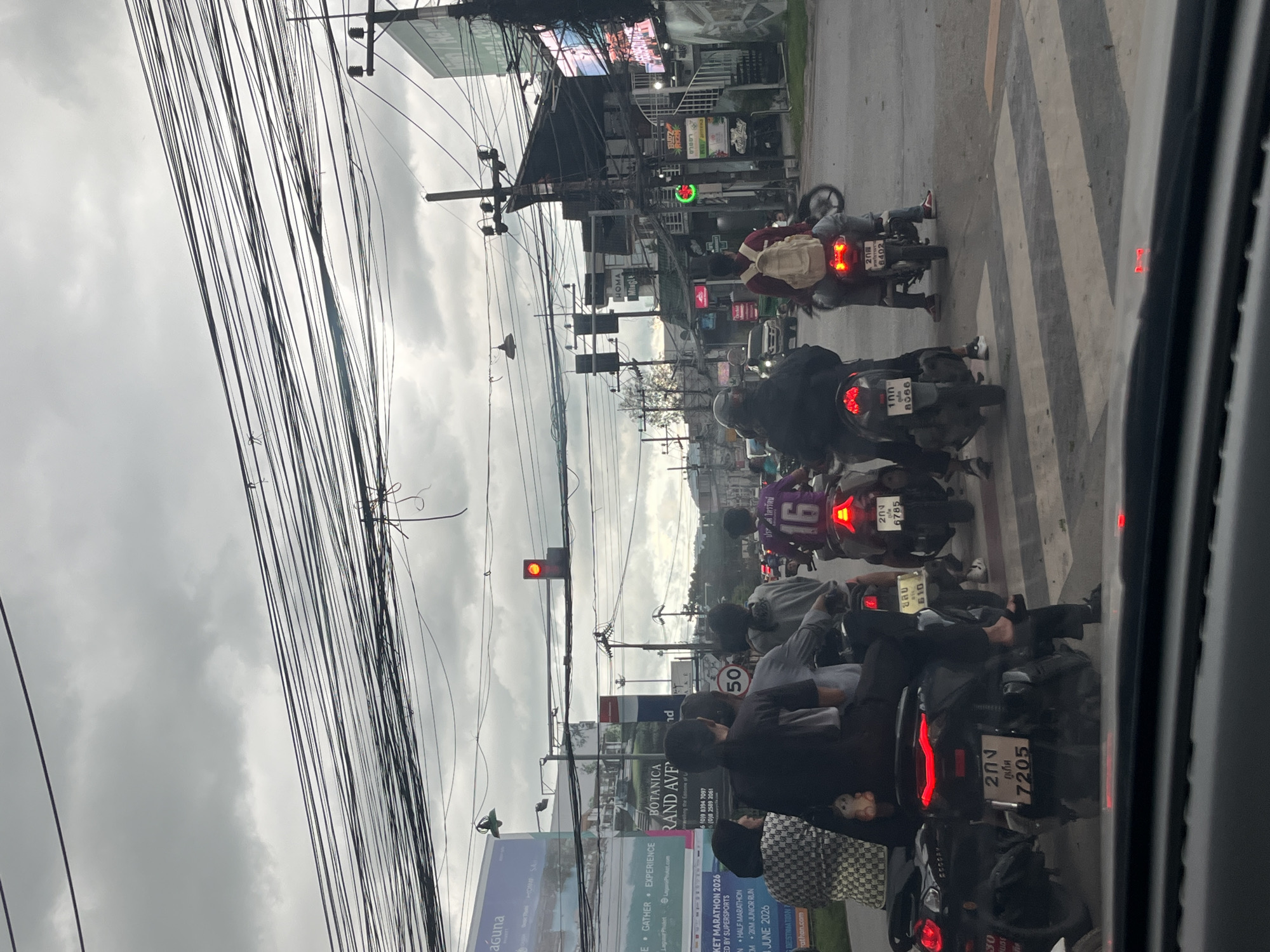 This image captures a traffic scene in Choeng Thale, Thailand, under a cloudy sky. A group of people on motorcycles are stopped at an intersection, likely at a crosswalk. The focus is on the motorcycles and their riders, with several vehicles in the foreground and middle ground. The motorcycles are varied in color and model, with some displaying license plates.
In the foreground, two motorcycles are visible. The one on the left is black with a red taillight and a license plate reading "100 8086". The rider is wearing dark clothing. The motorcycle to its right is also black, with a red taillight and a license plate with Thai characters and the number "7205". This rider is wearing dark pants and black footwear.
In the middle ground, several other motorcycles and riders are present. One rider in purple is wearing a jersey with the number "16" on the back. Another rider wears a light-colored backpack. Further back, a white car is stopped in traffic, and the general urban environment of Choeng Thale, with buildings and power lines, is visible. A traffic light is illuminated red in the distance, indicating a stop. The scene suggests a typical busy intersection in a Thai city.