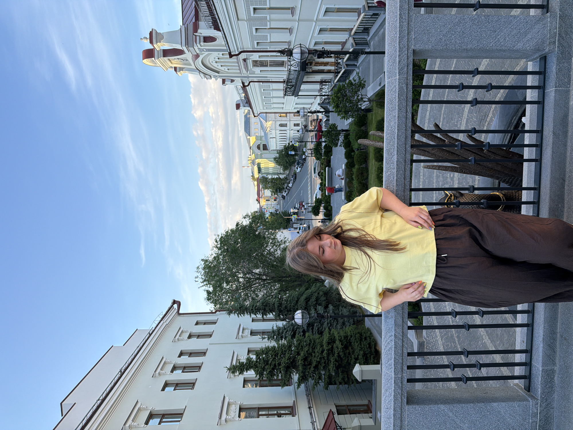 A young woman stands on an elevated gray granite platform, leaning on a railing with black vertical balusters, overlooking a city streetscape in Kazan, Russia. She has long, light brown hair and wears a loose-fitting pale yellow short-sleeved t-shirt and dark brown wide-leg pants. Her hands are placed together on her midsection, revealing brightly painted fingernails in various colors. Her gaze is directed downwards and slightly to her right, away from the camera.
To her left, a light-colored, multi-story building with multiple brown-framed windows and white decorative elements above them is partially obscured by several tall, green evergreen trees and a larger deciduous tree. A black lamppost with a spherical glass fixture stands in front of the trees. To her right, a more ornate, light-colored building with a distinctive red and white roofline, featuring curved gables and finials, stands prominently. This building includes a black wrought-iron balcony.
Below the elevated platform and extending into the background is a street, likely Bauman Street (Ulitsa Baumana) or a parallel historic street in central Kazan, lined with parked cars and a few moving vehicles. Pedestrians are visible on sidewalks. The street is flanked by further historic buildings with varying architectural details, receding into the distance. Below the railing and in the paved area, a large, dark bronze-colored sculpture, identifiable as a stylized claw or wing of the Zilant (Dragon) sculpture, is partially visible. The sky above is clear blue with scattered white, wispy clouds, suggesting a late afternoon or early evening setting.