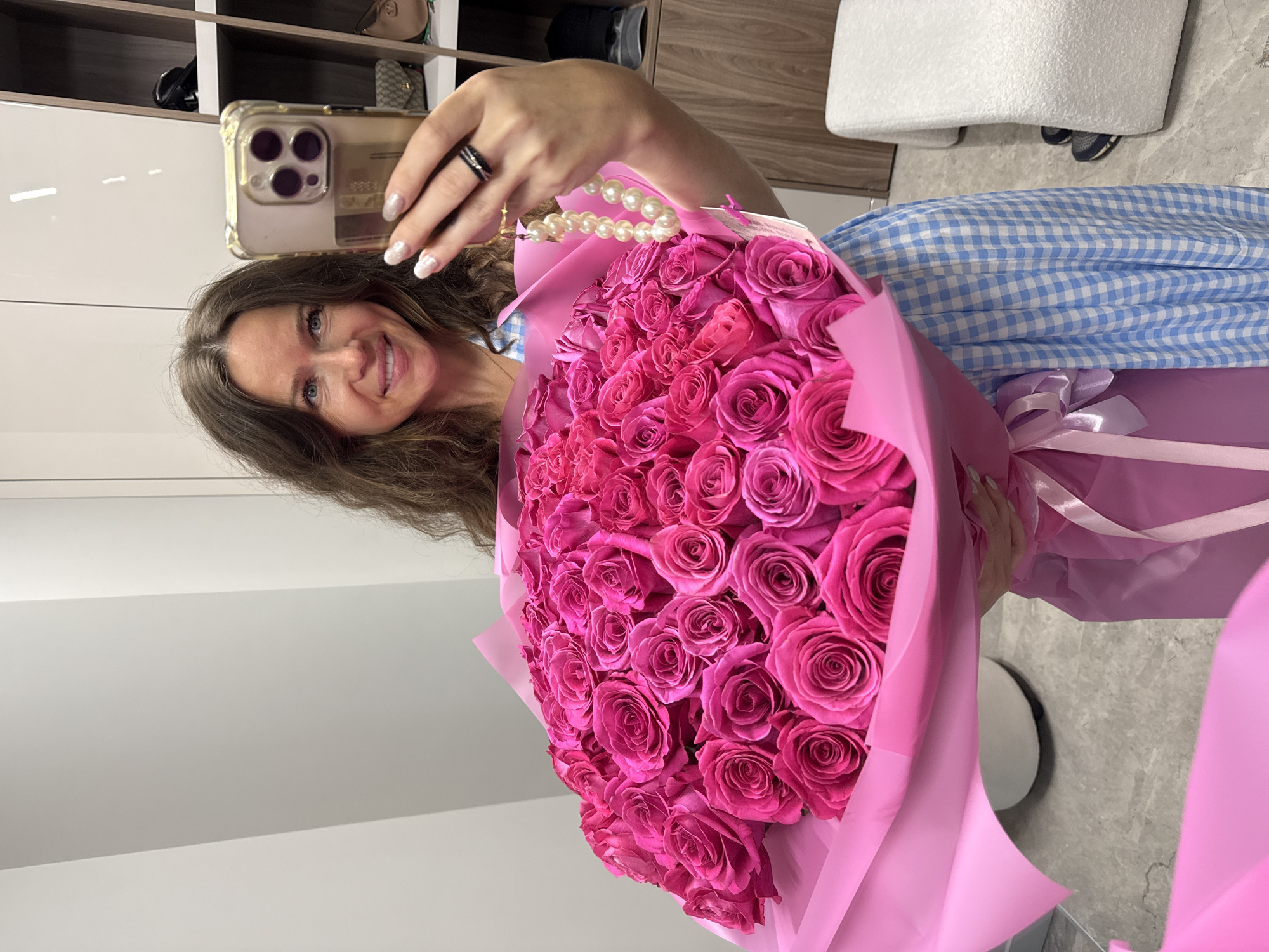 The image shows a woman taking a selfie with a large bouquet of pink roses. She is holding a smartphone in her left hand and the bouquet with her right hand. The roses are arranged in a dense cluster and wrapped in pink paper. The woman has long, wavy brown hair and is smiling at the camera. She is wearing a blue and white checkered dress. In the background, there are shelves with various items, including bags and what appear to be clothing items. The setting seems to be an indoor space, possibly a room or a walk-in closet. The lighting suggests it is daytime. The overall mood of the image is cheerful and celebratory.