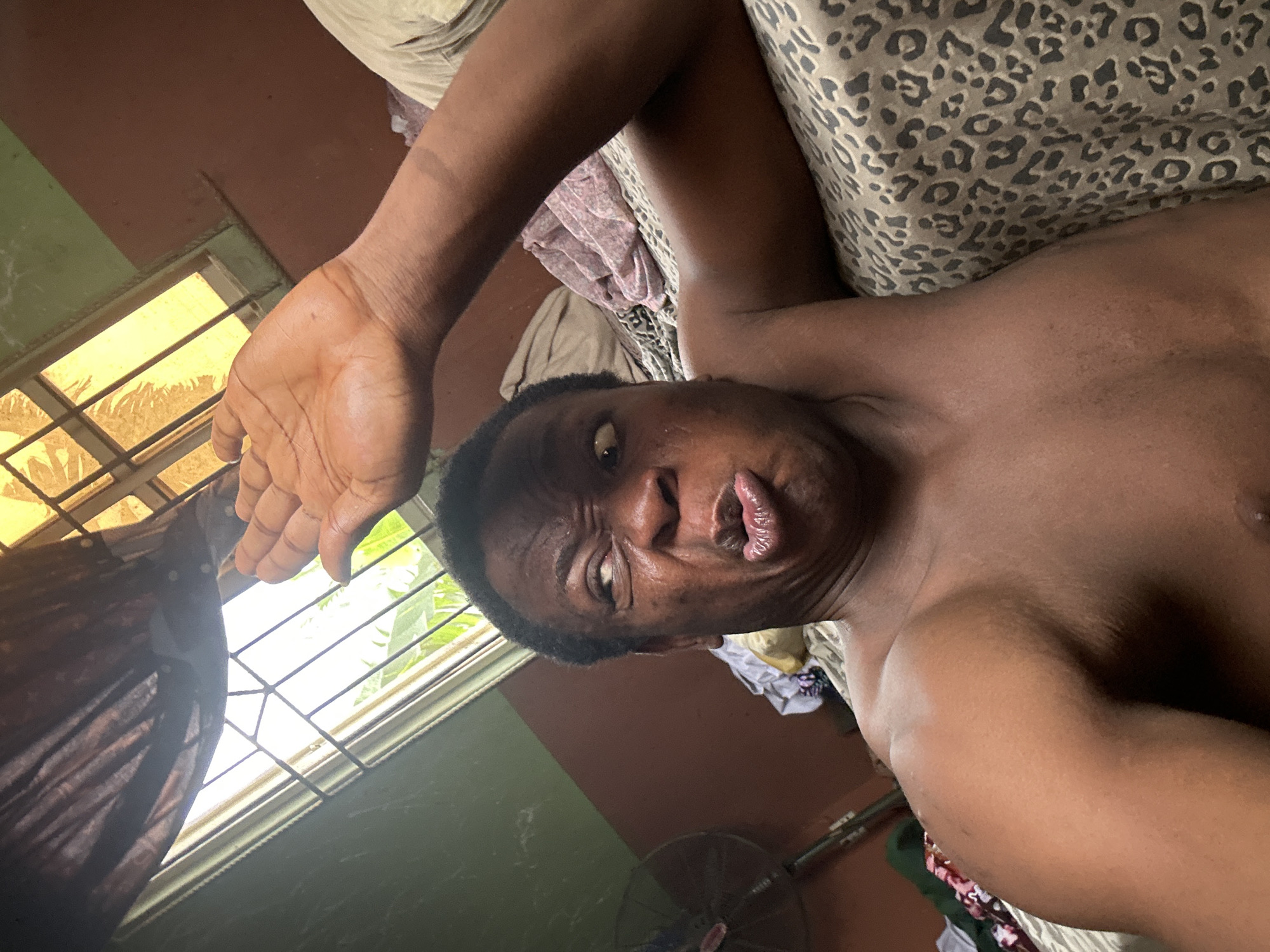 A shirtless dark-skinned person is lying on a bed with a leopard print patterned blanket. The person is positioned at an angle, looking directly at the camera with a slightly contorted facial expression. Their left arm is raised, with their hand held up near a window with bars. Outside the window, greenery is visible. The room is painted in two tones: brown and green. In the lower right corner of the image, a black fan can be seen.