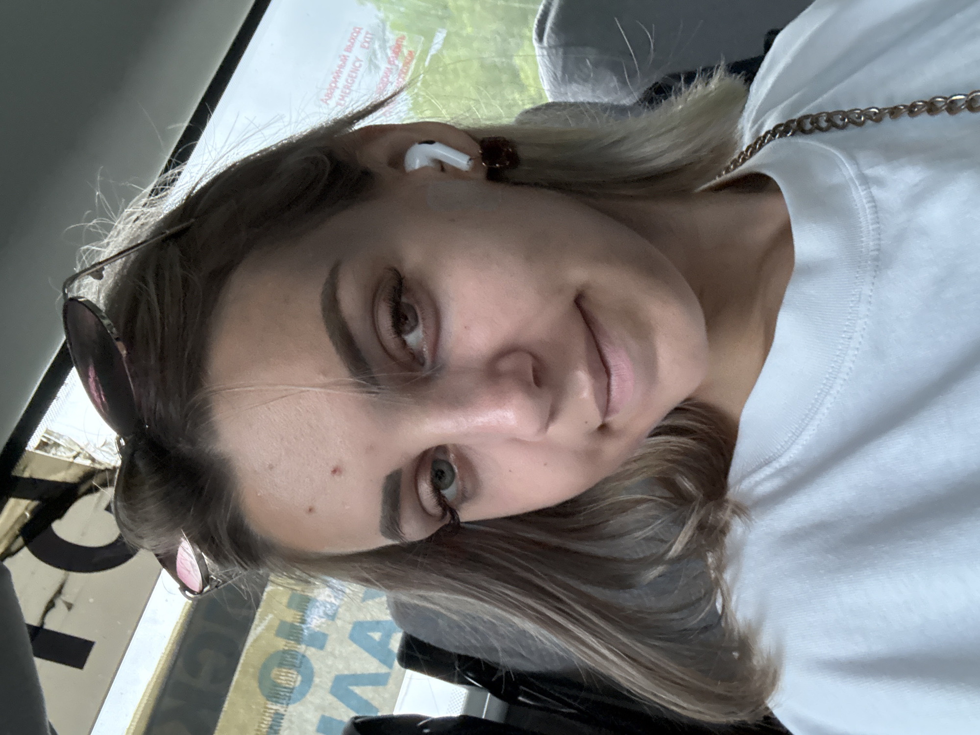 A young woman with blonde hair is taking a selfie in what appears to be the interior of a bus or train. She is wearing a white t-shirt and has on white wireless earbuds in her right ear, with a dark red earring. Sunglasses are perched on top of her head. The background visible through the window suggests a green, treed environment, consistent with a location outside of Ogon'ki, Russia, where the bus or train may be traveling. A partial sign with Russian text, "Аварийный выход EMERGENCY EXIT", is visible on the window.