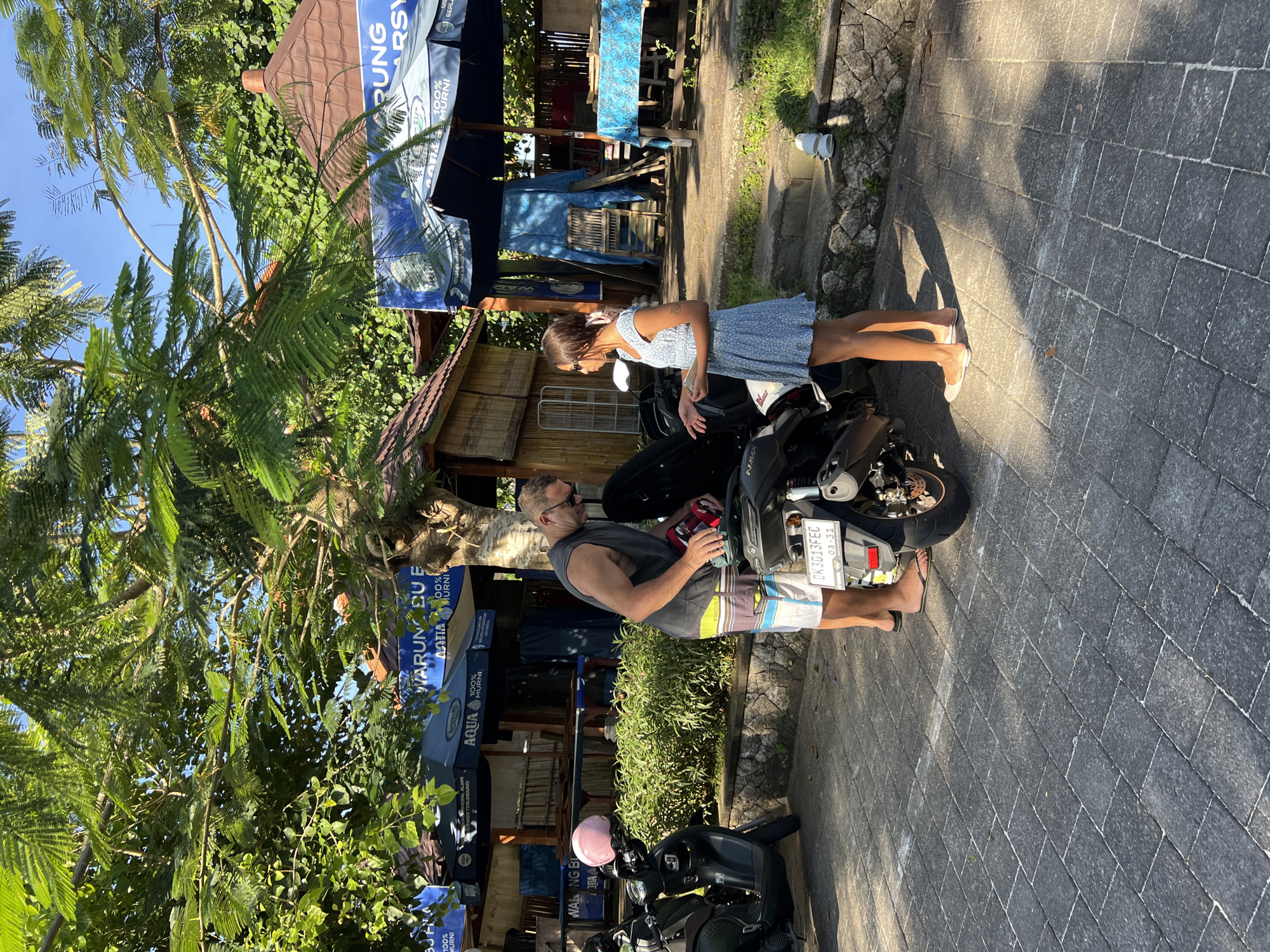 Observed in an outdoor scene in the city of Unknown, Unknown, are two individuals positioned near two black motorcycles. A man, wearing sunglasses, a gray tank top, and multi-colored shorts, is standing by a motorcycle and appears to be holding an item. Next to him, a woman in a short, light-blue floral dress is leaning over the other motorcycle. Visible on the motorcycle is a license plate with the designation "DK3013FEC". Overhead, there's an umbrella with text containing "WARUNG EU" and "AQUA" printed on it. Lush greenery, including trees, surrounds the scene, and a building with a bamboo exterior is in the background. The ground is paved with interlocking pavers.