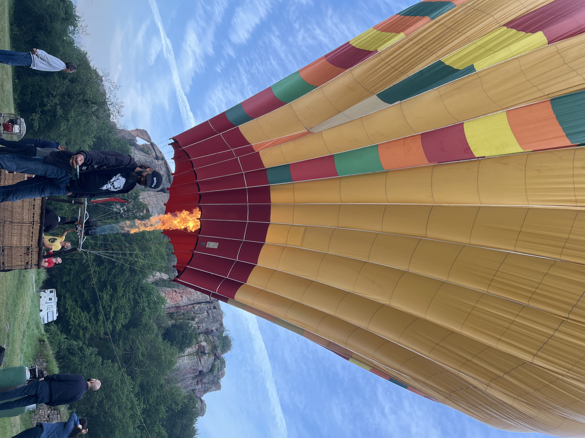 A large, multi-paneled hot air balloon is in the process of inflation on a grassy field, viewed from a low angle looking upwards. The balloon's envelope is predominantly golden-yellow, with horizontal bands of red, orange, green, and lighter yellow-brown fabric visible on its lower section. The very bottom section, or skirt, of the balloon is a deep red. A powerful flame, bright orange and yellow, emanates from the burner system directly beneath the balloon's opening, indicating active heating.
Several individuals are present on the ground. In the immediate foreground, slightly to the left of center, a man with a dark beard, wearing a black baseball cap with "Motorhead" text, a black t-shirt with a white skull graphic, a black jacket, and blue jeans, holds a small electronic device in his right hand. To his left, another man in a white long-sleeved shirt and blue jeans stands with his hands behind his back, looking upwards towards the balloon. Near the wicker basket, a man in a yellow t-shirt is actively attending to the burner system, looking up at the flame. To the right of the basket, two other individuals observe the scene; one in a dark blue shirt and dark trousers, and another partially visible with dark, curly hair. A red and silver industrial fan is positioned on the grass to the left of the basket, and two green propane tanks are visible to the right.
The background features towering, reddish-brown rock formations with eroded surfaces and sparse green vegetation, indicative of a rugged natural landscape. Numerous green trees and bushes surround the field and dot the base of the rock formations. A white camper van is parked further in the middle ground, partially obscured by trees. The sky is a light blue with scattered white, wispy clouds, and a faint contrail is visible in the upper right. The scene takes place in the vicinity of Unknown, Unknown.
