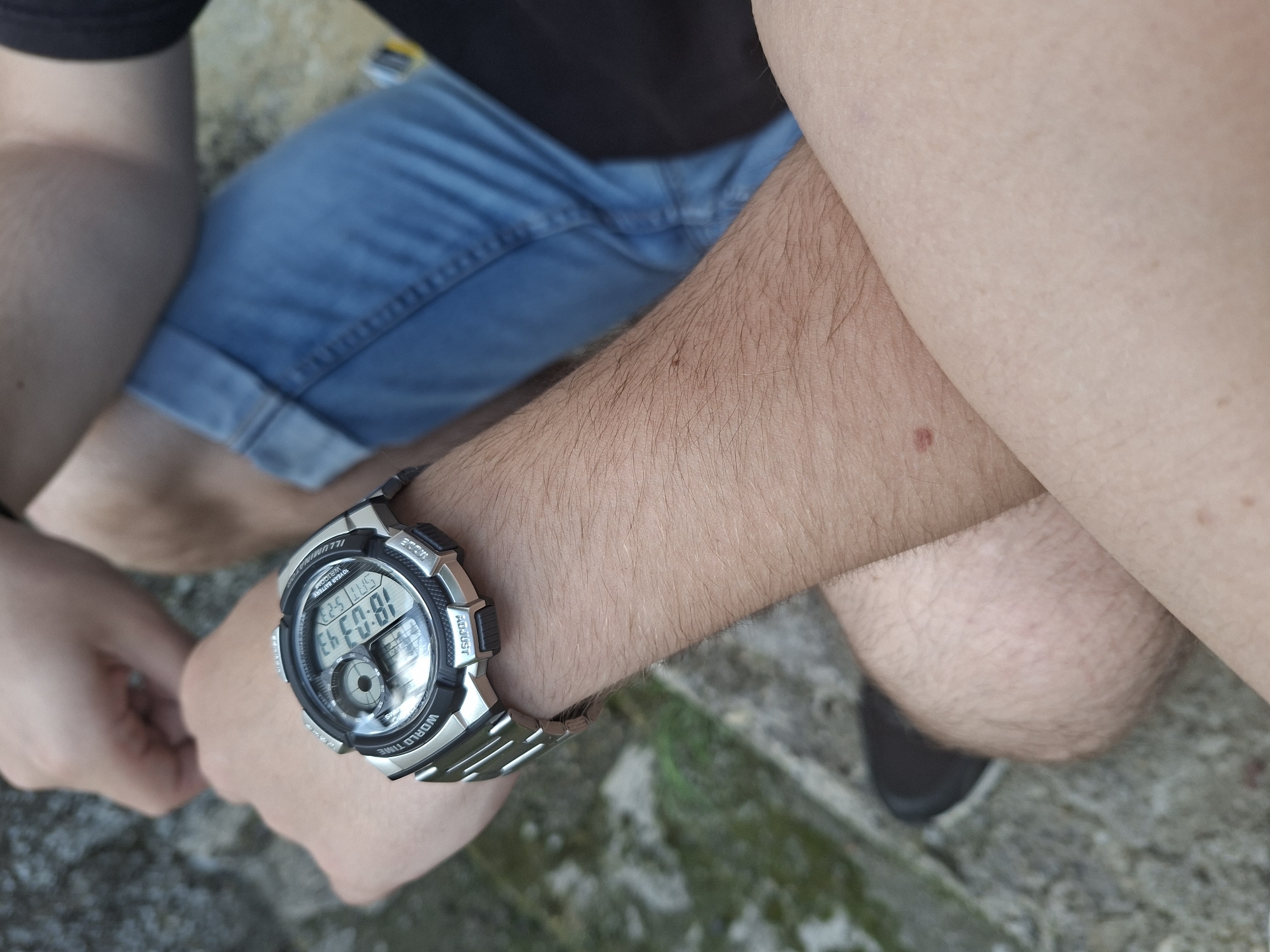A person is sitting with their arm resting on their leg. They are wearing blue jean shorts and a black shirt. The person is wearing a digital watch on their left wrist that displays 18:03. The location appears to be outdoors, possibly on a concrete step or ledge. The background is blurry and indistinct, making it impossible to verify the Svirachi, Bulgaria location.