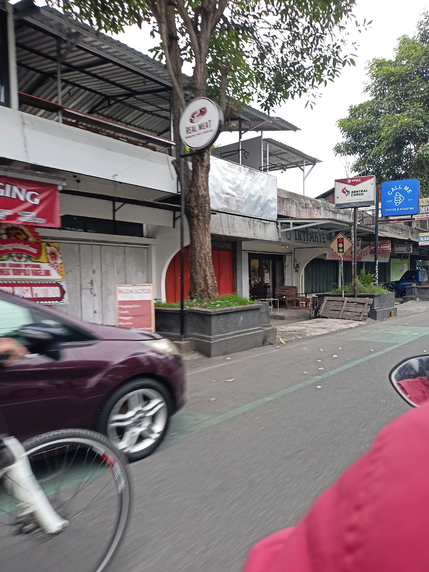 The image shows a street scene in Magelang, Indonesia, with a focus on commercial establishments. On the left, a red banner with white lettering, partially obscured, is visible. Adjacent to it, a maroon car is in motion, with its front wheel and part of its body in the foreground. Behind the car, a building with a white facade and metal awnings is seen, featuring several closed doors. A circular sign with "REAL MEAT" is mounted on a pole next to the building, beneath a tree. Further down the street, more commercial signs are displayed, including a blue sign that reads "CALL ME COFFEE & ROASTER". Several other businesses with awnings and signs line the street. The environment appears to be a sidewalk with planters and some debris on the road. The time of day seems to be daytime, with overcast or bright conditions indicated by the lighting.