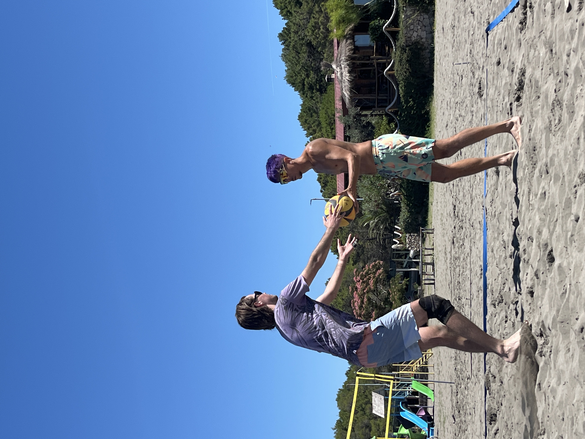 Two young men are playing beach volleyball on a sandy court under a clear blue sky. The man on the left, wearing a purple patterned t-shirt, light blue and pink shorts, sunglasses, and a black knee brace, is in the process of passing a volleyball to the man on the right. The man on the right has purple hair, is wearing sunglasses, and patterned shorts. He is holding the volleyball with both hands, preparing to receive the pass. In the background, there is a children's playground with slides, a beach volleyball net, trees, and a rustic beach hut. The sand is dark and appears to be a typical beach setting. The image captures a moment of active recreation on a sunny day.