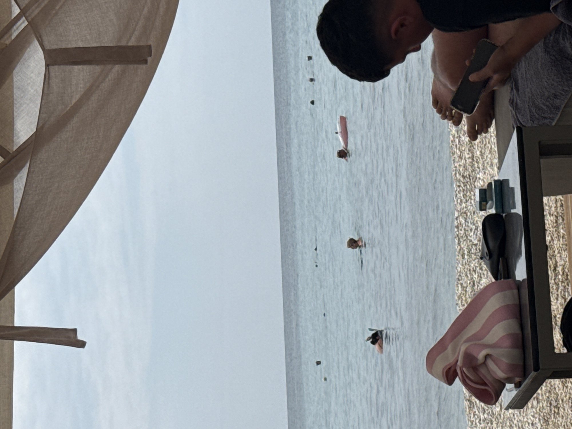 A view from beneath a fabric awning at a beach resort in Limenaria, Greece. The sea is calm and a light blue, with a few people swimming or snorkeling in the distance. In the foreground, a man is sitting on a lounge chair, looking at his phone. A striped beach bag, a pair of black flip-flops, and a small stack of what appear to be cigarette packs are on a table next to him. The beach is covered in small, light-colored pebbles. The sky is overcast.