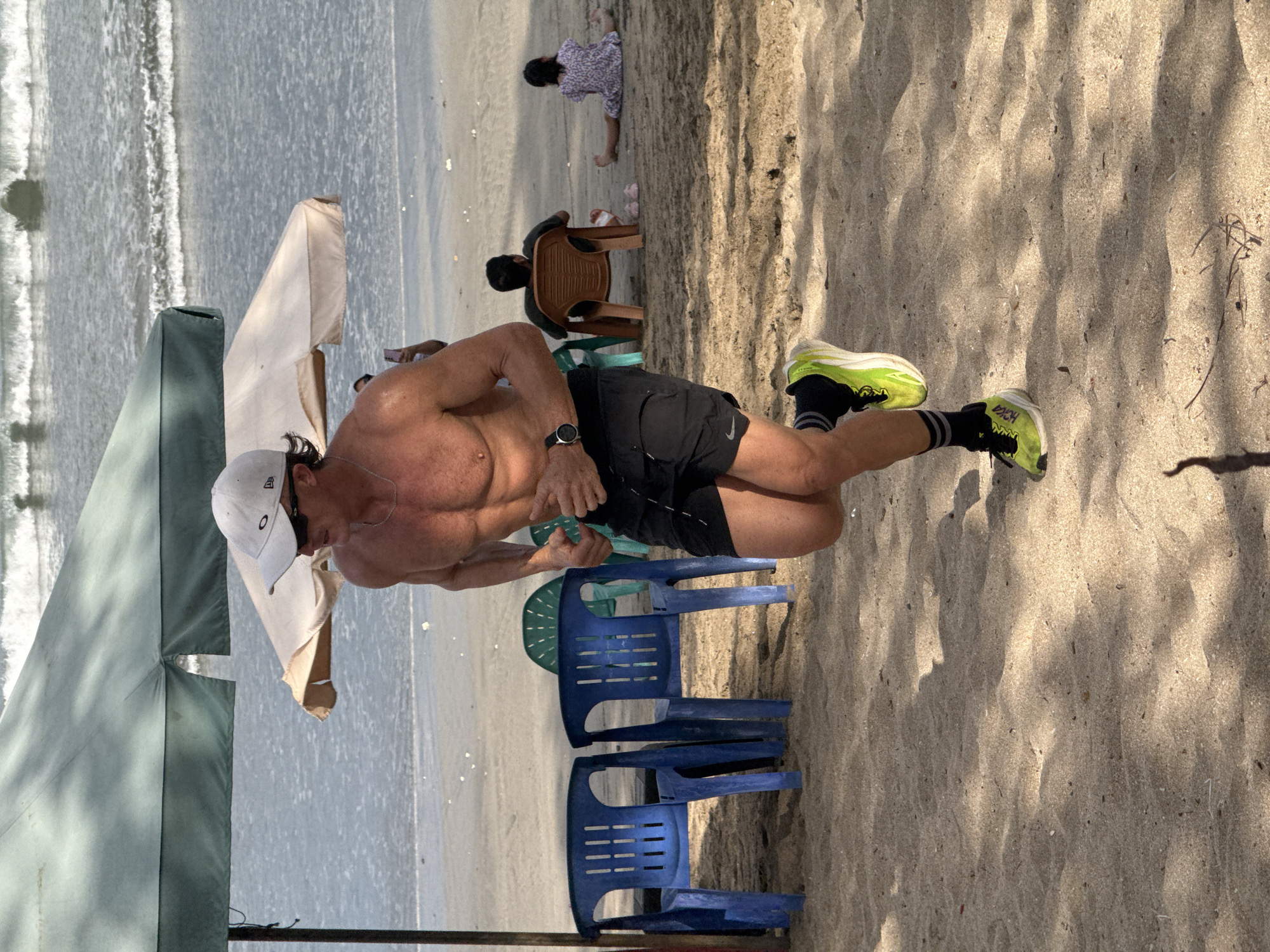 A shirtless man with muscular physique is jogging on a sandy beach in Kuta, Indonesia. He is wearing black athletic shorts, a white baseball cap, sunglasses, and bright lime-green running shoes with black socks. He has a watch on his left wrist. Behind him, there are two blue plastic chairs, partially shaded by a large green umbrella. Further back, other beachgoers are visible, including a person sitting in an orange chair and a child sitting on the sand. The ocean with gentle waves is in the background. The sun is casting shadows on the sand.
