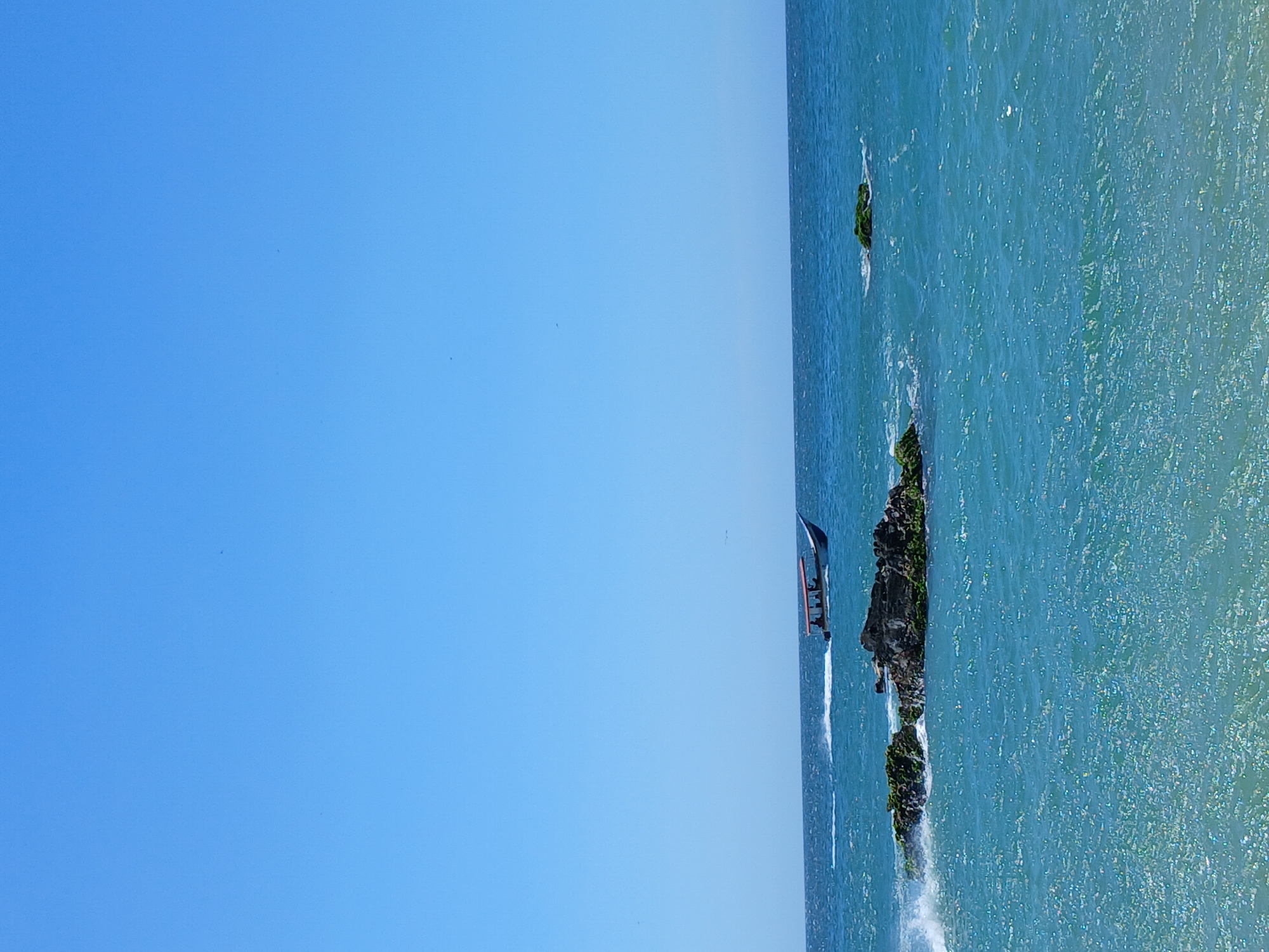 A vibrant coastal landscape, oriented horizontally, displays a clear blue sky dominating the left two-thirds of the frame and a sparkling turquoise ocean on the right. The sky is completely cloudless and bright.
The ocean's surface is rippled and shimmers with sunlight. Scattered within the water, particularly towards the center-right, are several dark rocky outcroppings, covered with green moss or algae. White foam and small waves crash against the bases of these rocks.
In the mid-ground, a small motorboat with several people on board travels across the water, moving towards the right side of the frame. The boat leaves a distinct white wake trailing behind it.
The scene depicts a sunny, clear-weather day, likely mid-day, consistent with a tropical or subtropical location like Pedro González, Venezuela. The overall impression is one of tranquility and natural beauty. There is no visible text in the image.