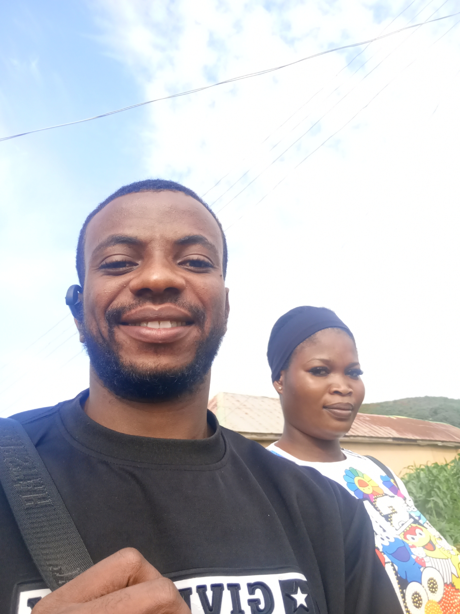 A medium shot captures a smiling Black man and a woman looking towards the camera. The man, in the foreground, wears a black t-shirt and a black earbud. The woman, positioned behind and to his right, wears a black headscarf and a white t-shirt with a colorful cartoon print. They appear to be outdoors, with a bright sky and some clouds in the background. A building with a reddish-brown roof and some greenery are visible behind them, suggesting a rural or semi-urban setting. The overall impression is of a casual, sunny day.